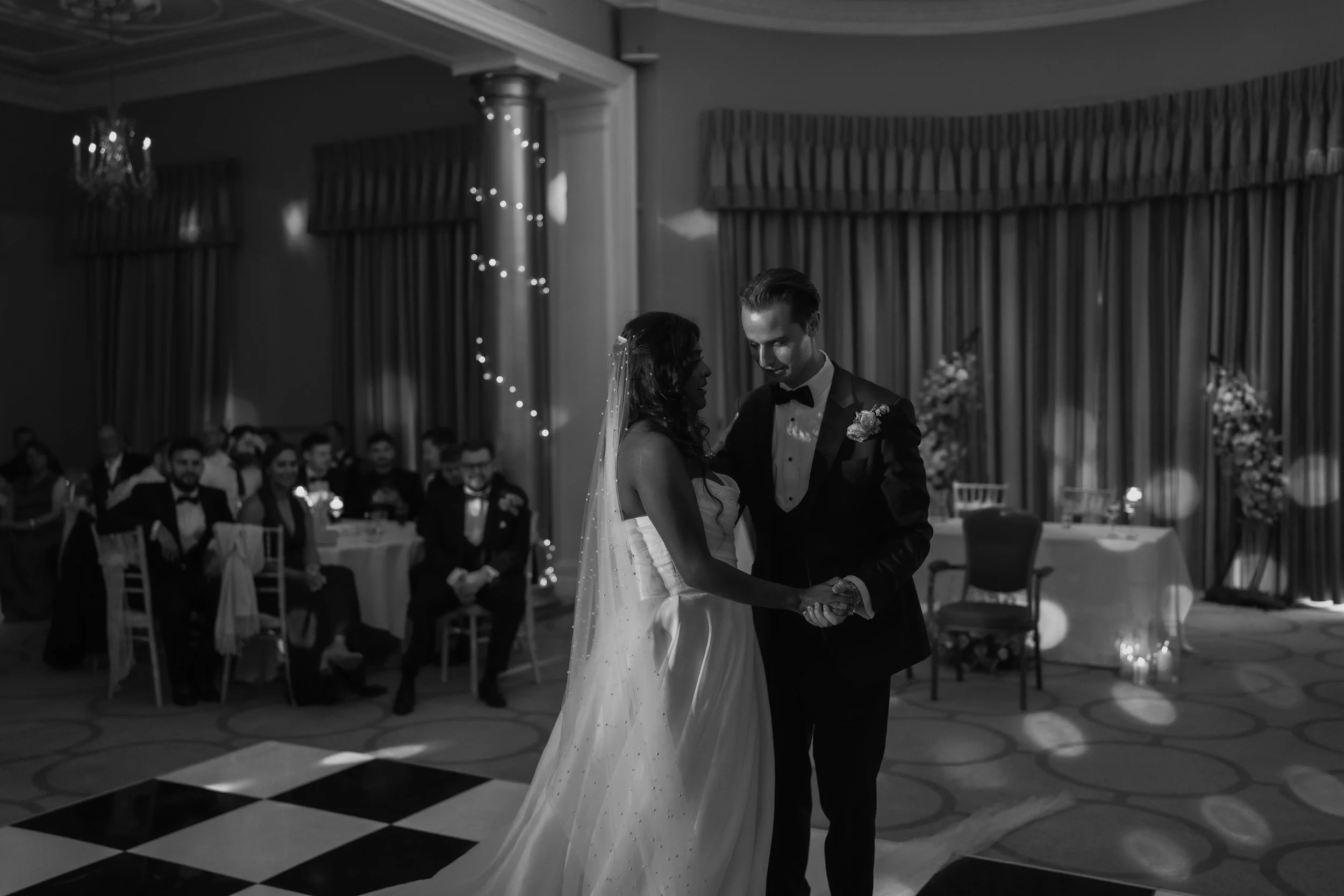 Black and white photo of a bride and groom sharing their first dance at their wedding reception, with guests seated in the background.
