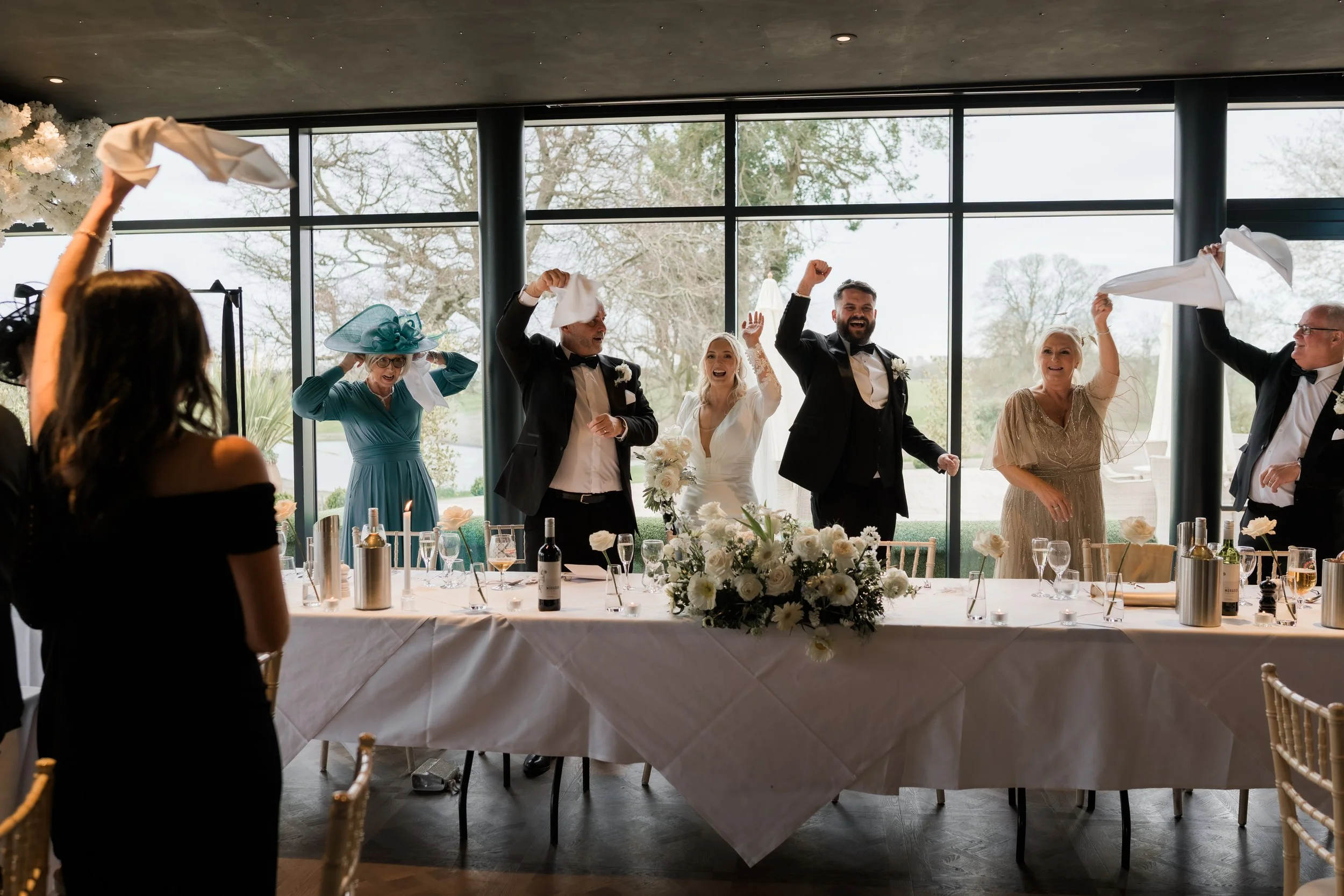 People celebrating at a wedding reception, holding napkins in the air, with a long decorated table and large windows showing a view of trees outside.