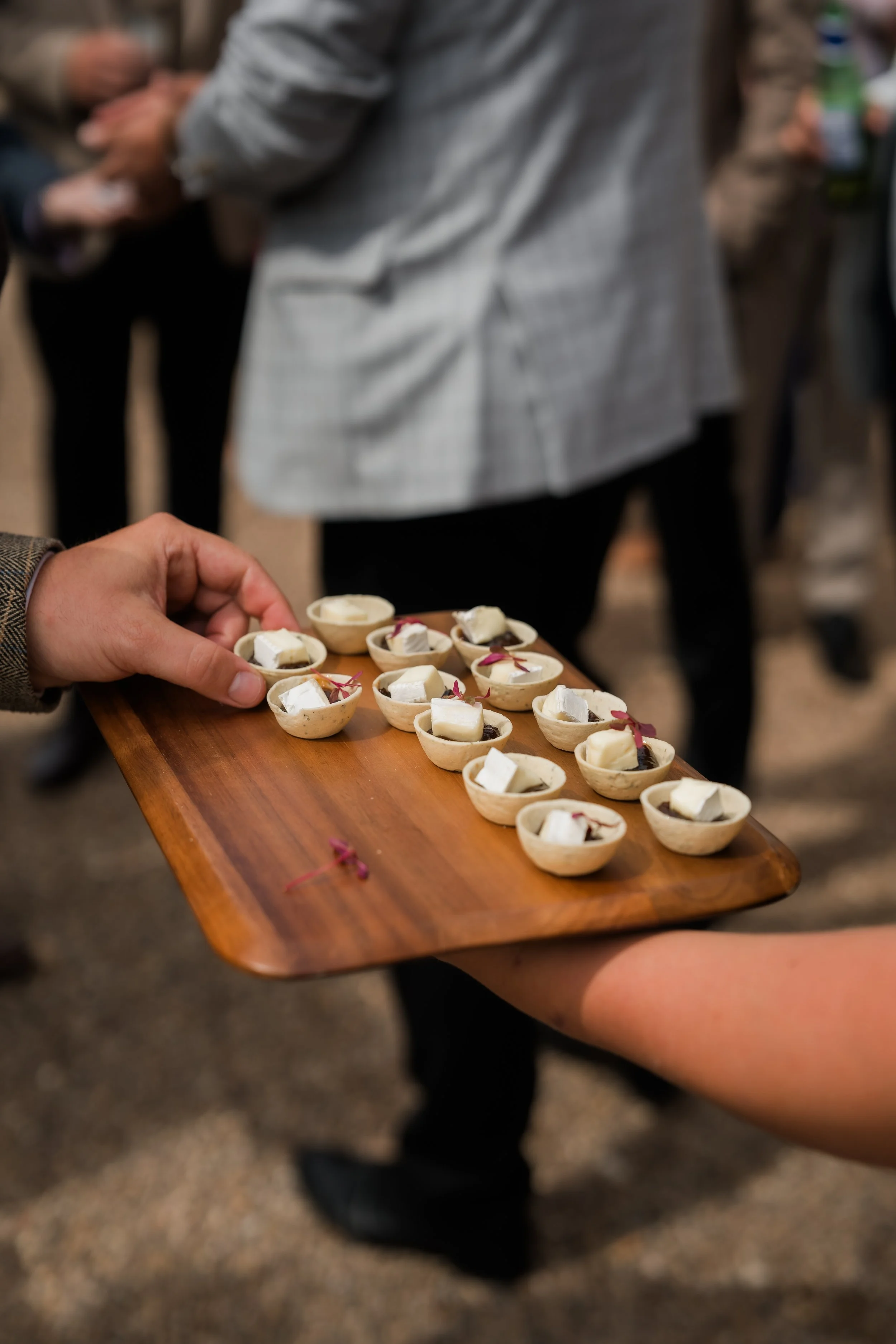 A person holds a wooden tray with small bowls of cheese and garnishes at an event.