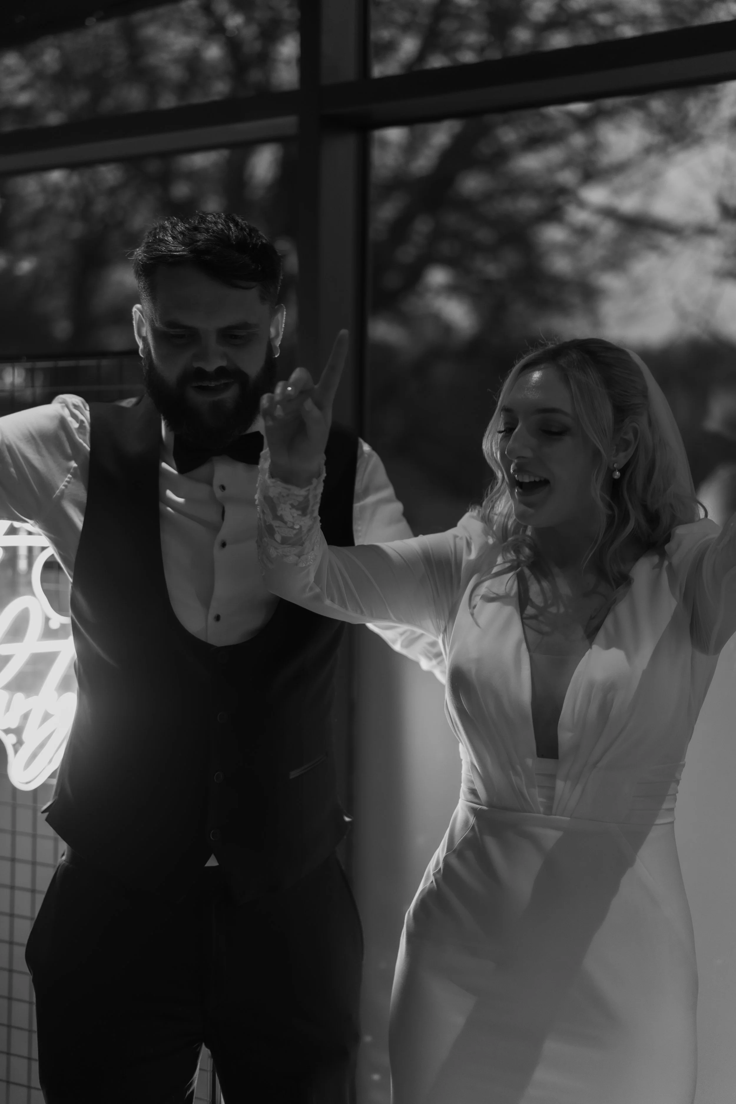 A black and white photo of a man and woman dancing together indoors, with the man wearing a tuxedo and the woman in a white dress, both smiling and enjoying the moment.