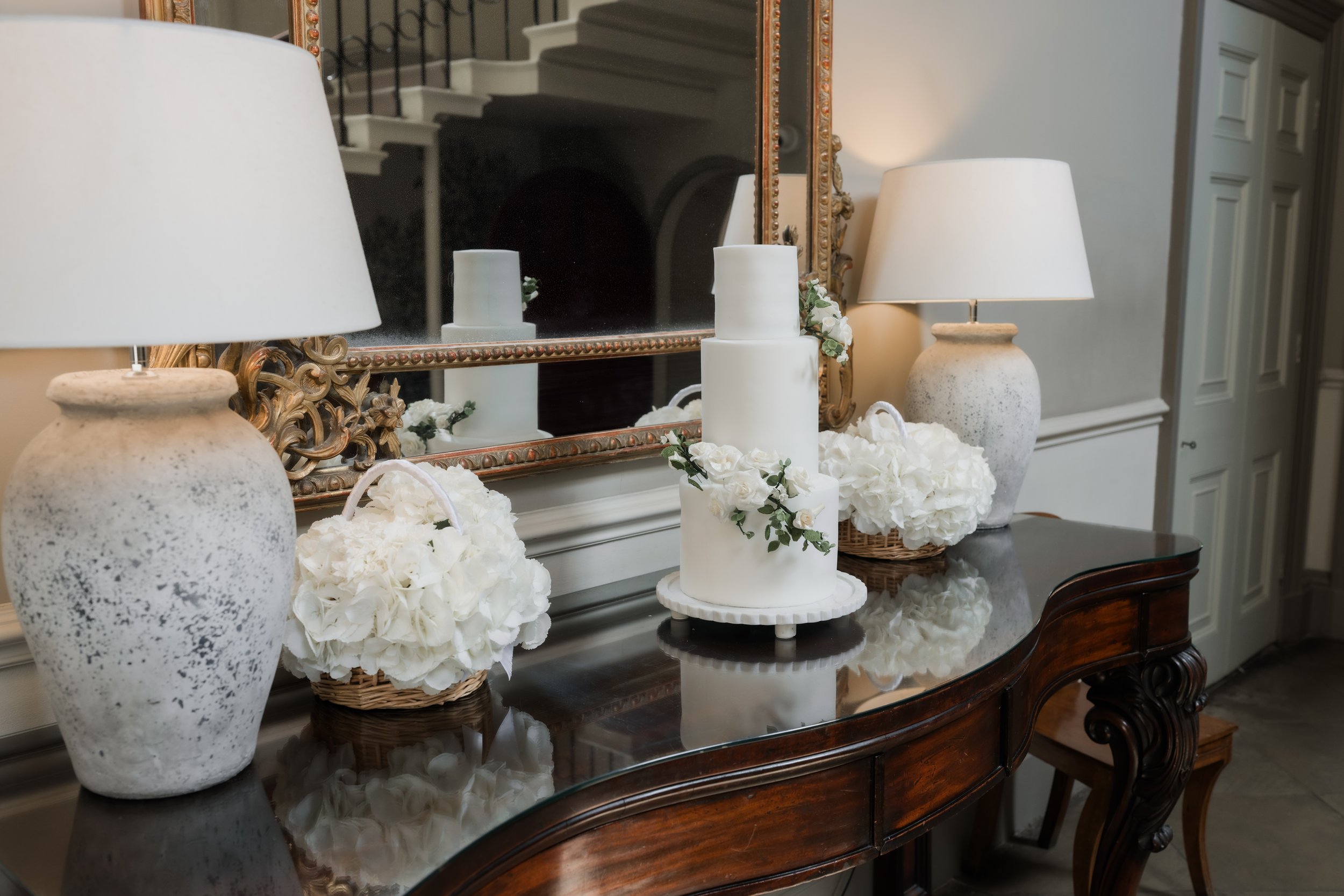A decorated mantelpiece with white candles, floral arrangements, and lamps, reflected in a mirror in an elegant living room.