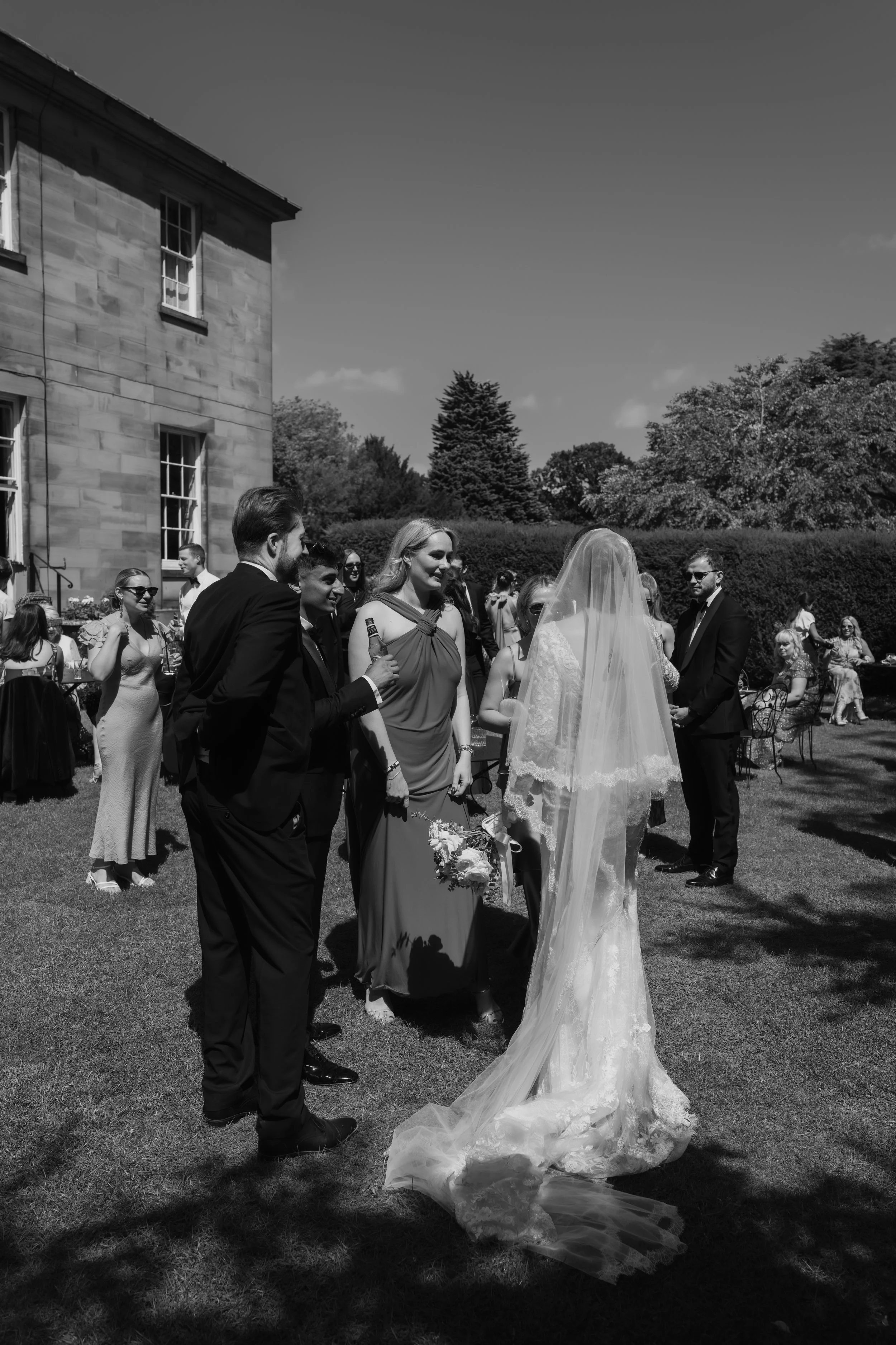 A wedding party outdoors on a sunny day, featuring a bride in a lace wedding gown with a long veil, holding a bouquet, and surrounded by guests dressed in formal attire.