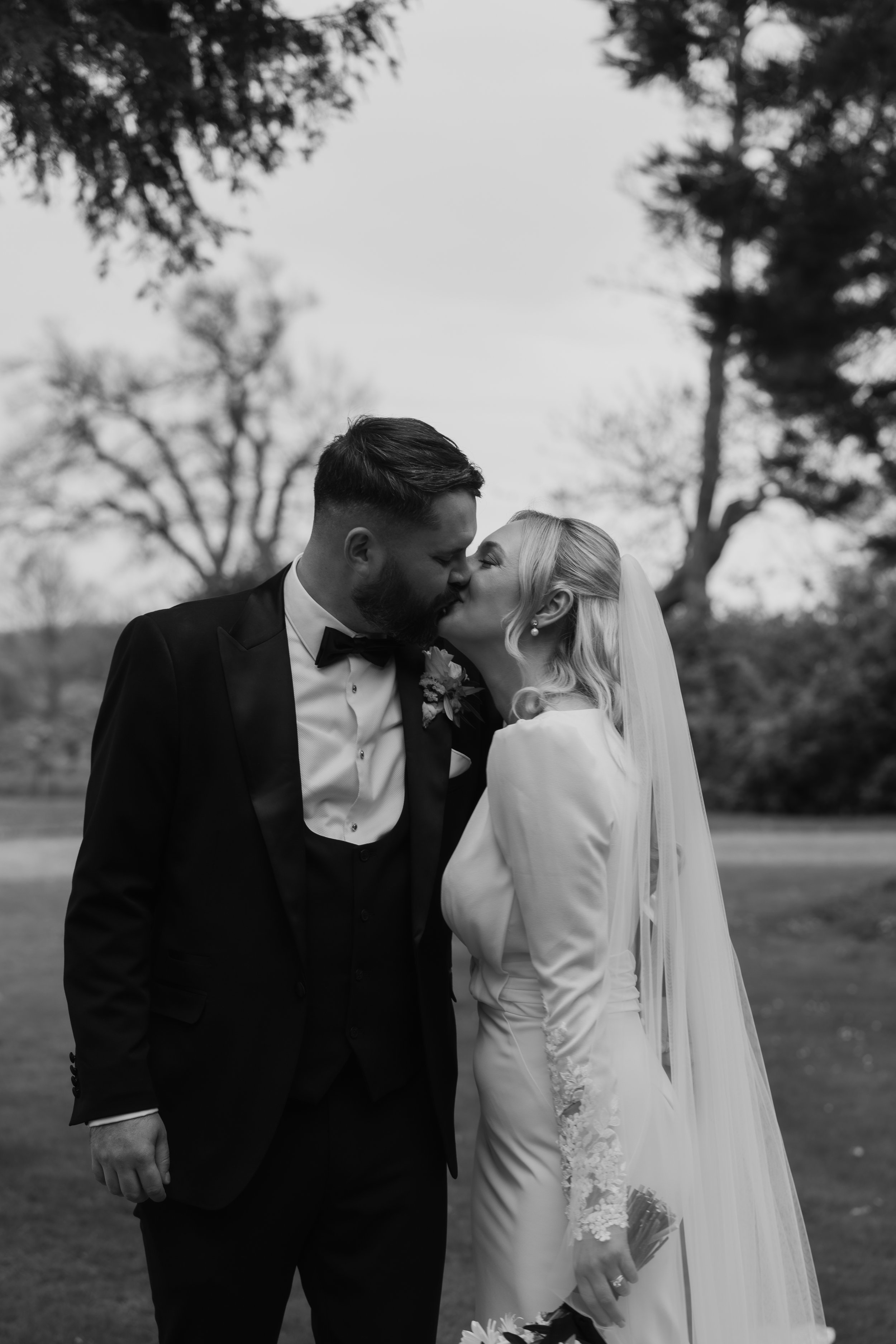 A black and white photo of a bride and groom kissing outdoors on their wedding day, with trees in the background.