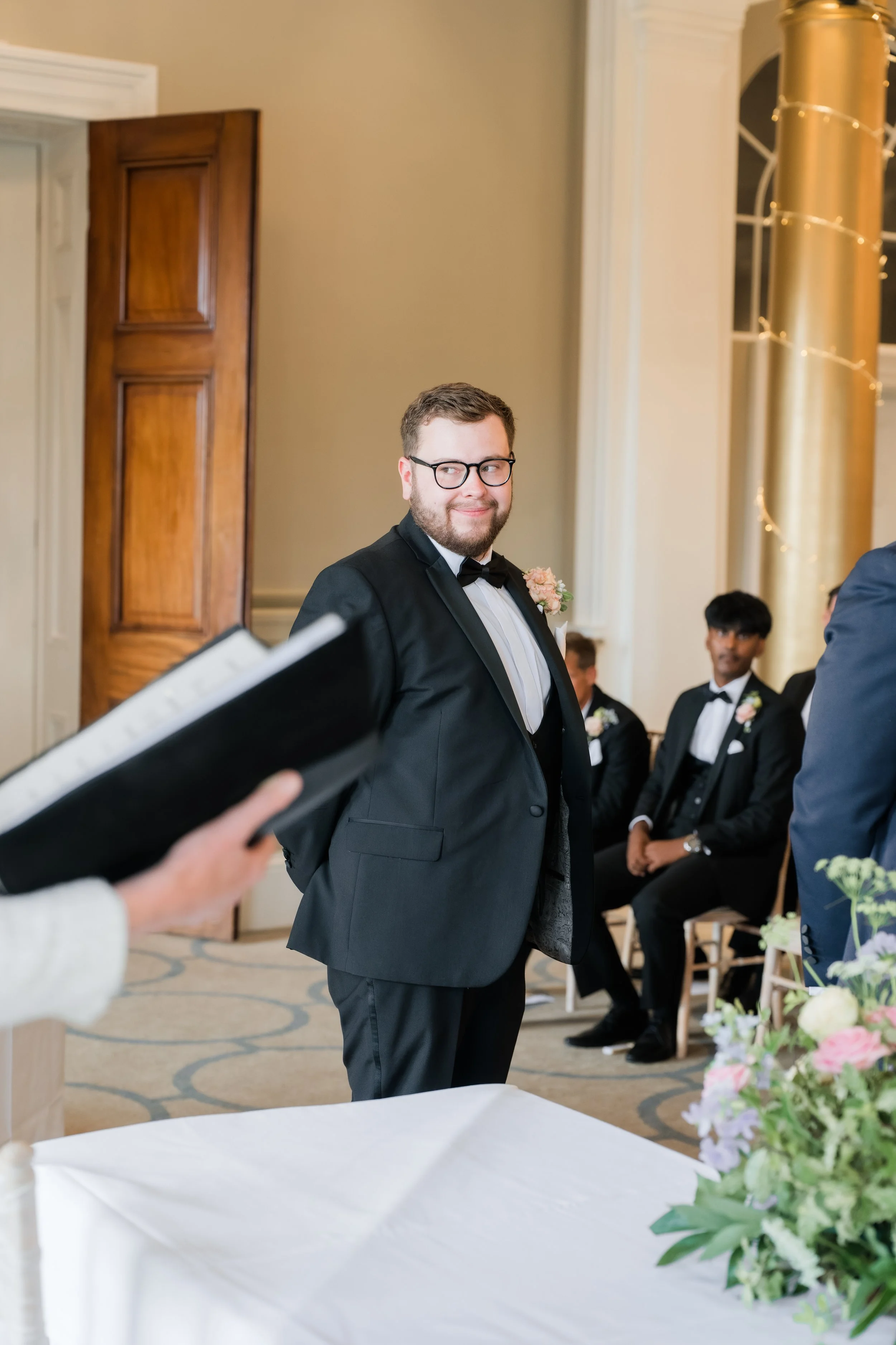 A groom in a black tuxedo and glasses at a wedding ceremony, standing in front of seated guests, with a flower boutonniere and a slight smile.