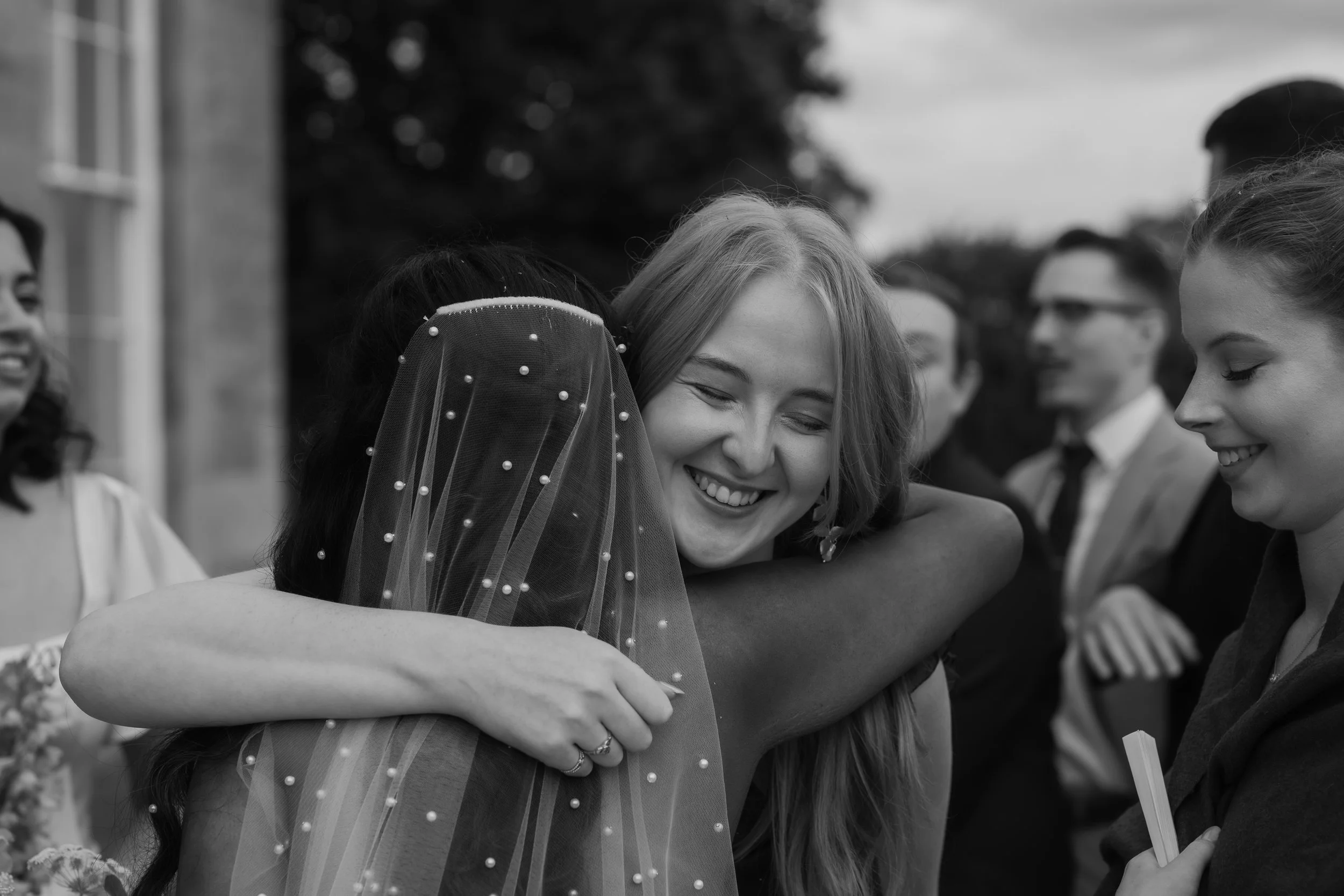 Two women hugging, one wearing a bridal veil, at a wedding celebration, smiling.