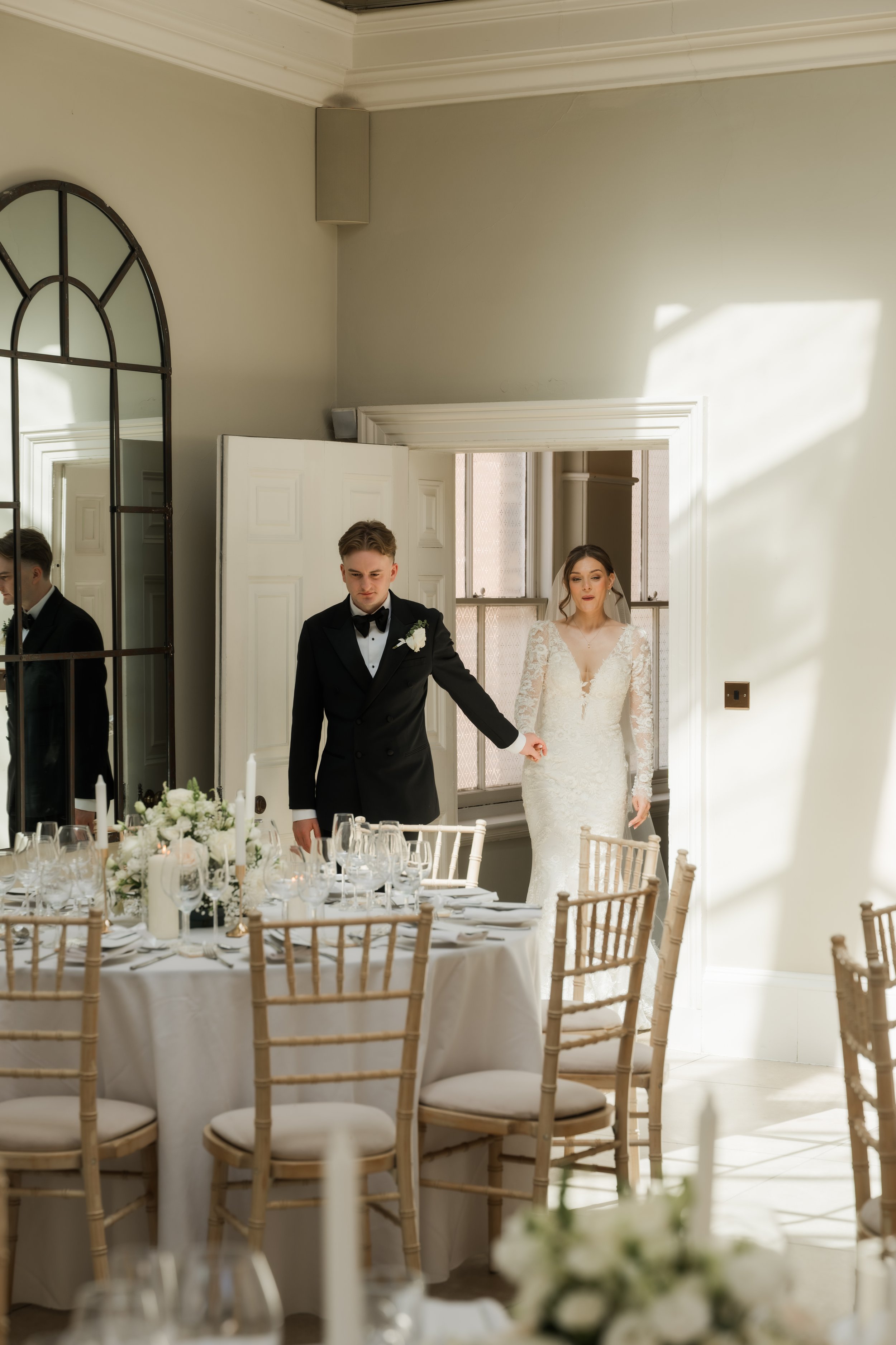 A bride and groom standing hand in hand in a softly lit room with round tables set for a wedding reception. The bride is wearing a lace wedding dress, and the groom is in a black tuxedo. The room features a large mirror, a window, and elegant decor.