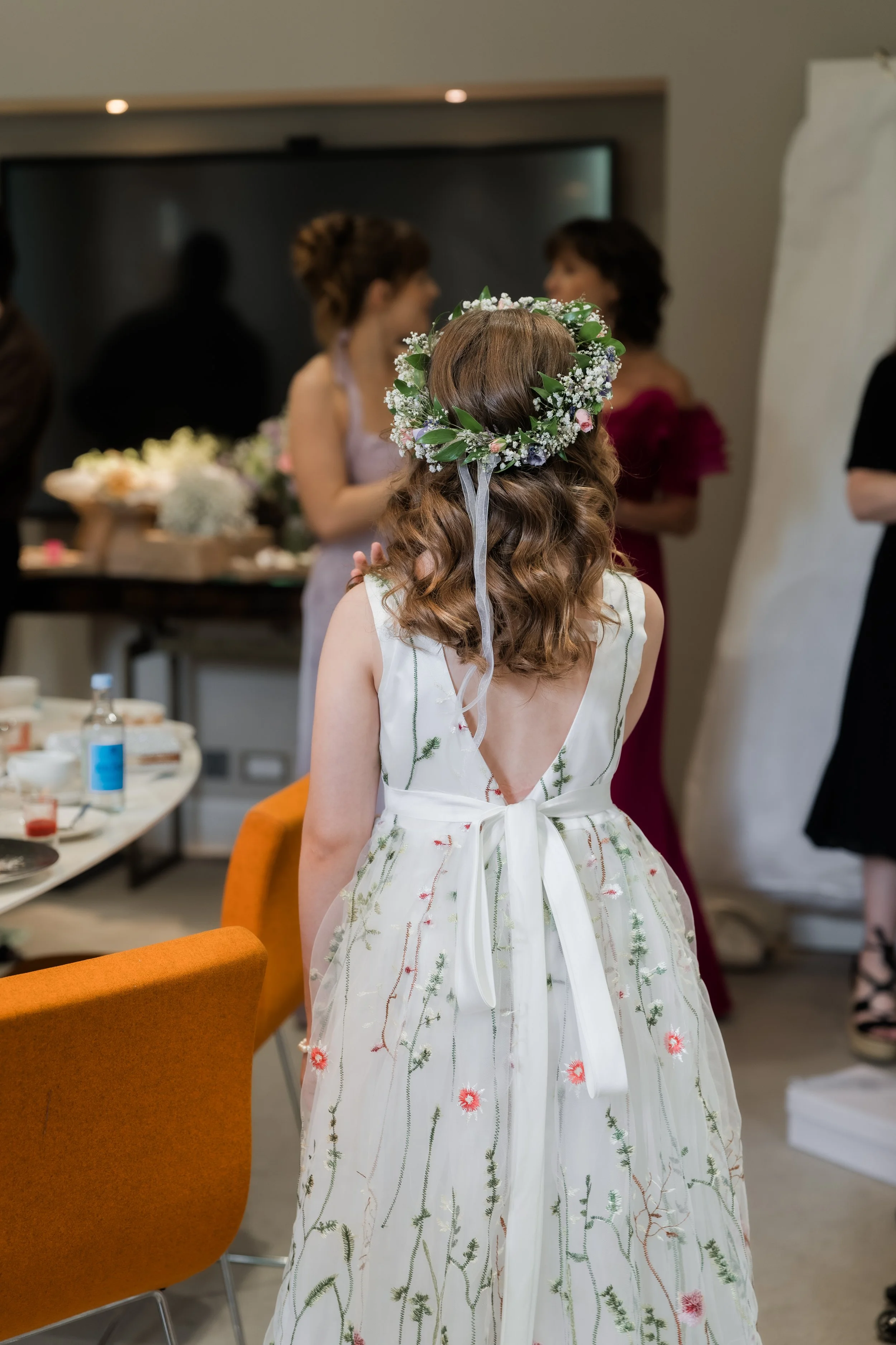 A woman with curly hair wearing a floral dress with an open back and a ribbon tie, and a flower crown, is at a gathering with other women in the background, some holding flowers.