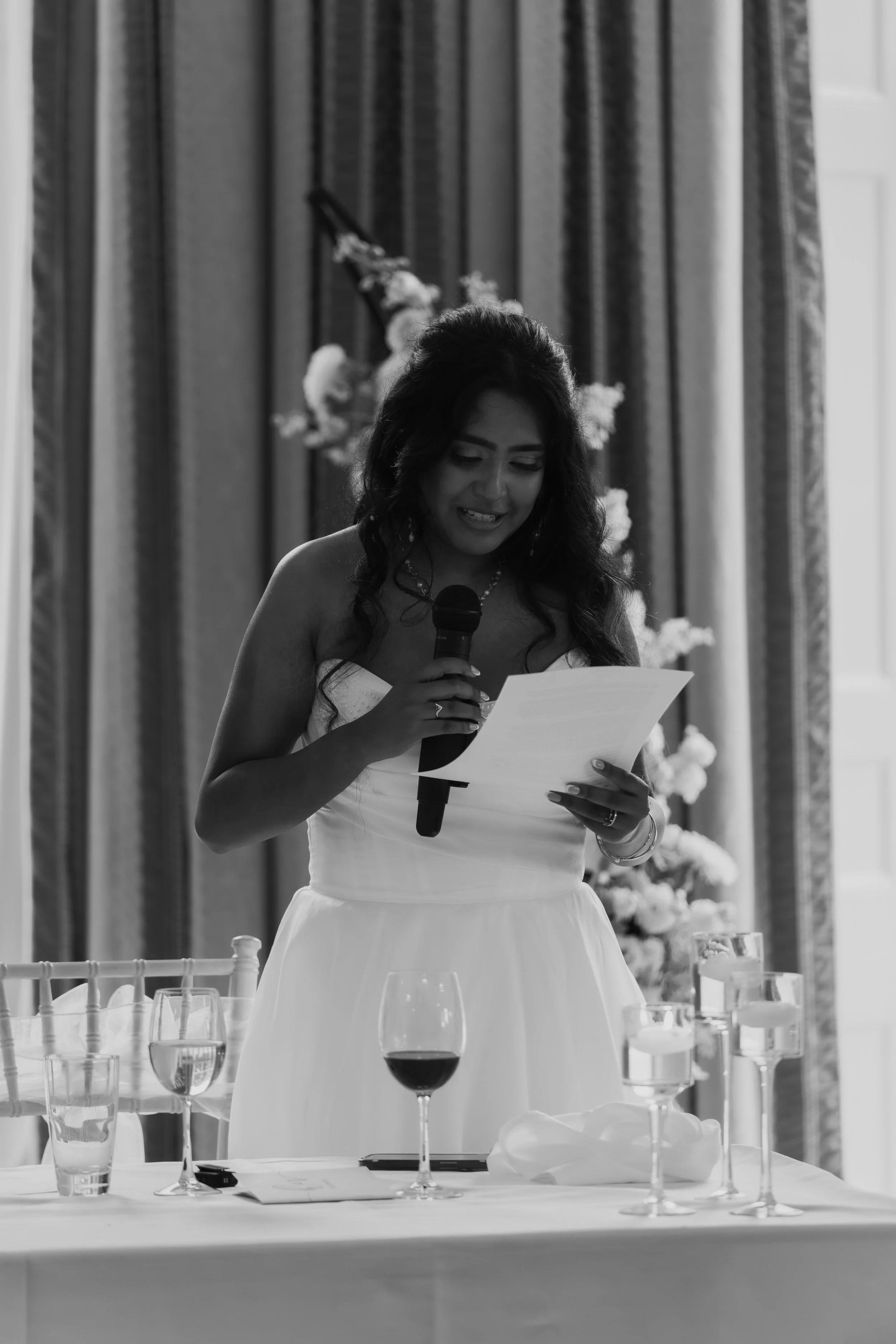 A woman in a white dress giving a speech at a wedding reception, holding a microphone and reading from a paper, with a table of drinks and a floral backdrop behind her.