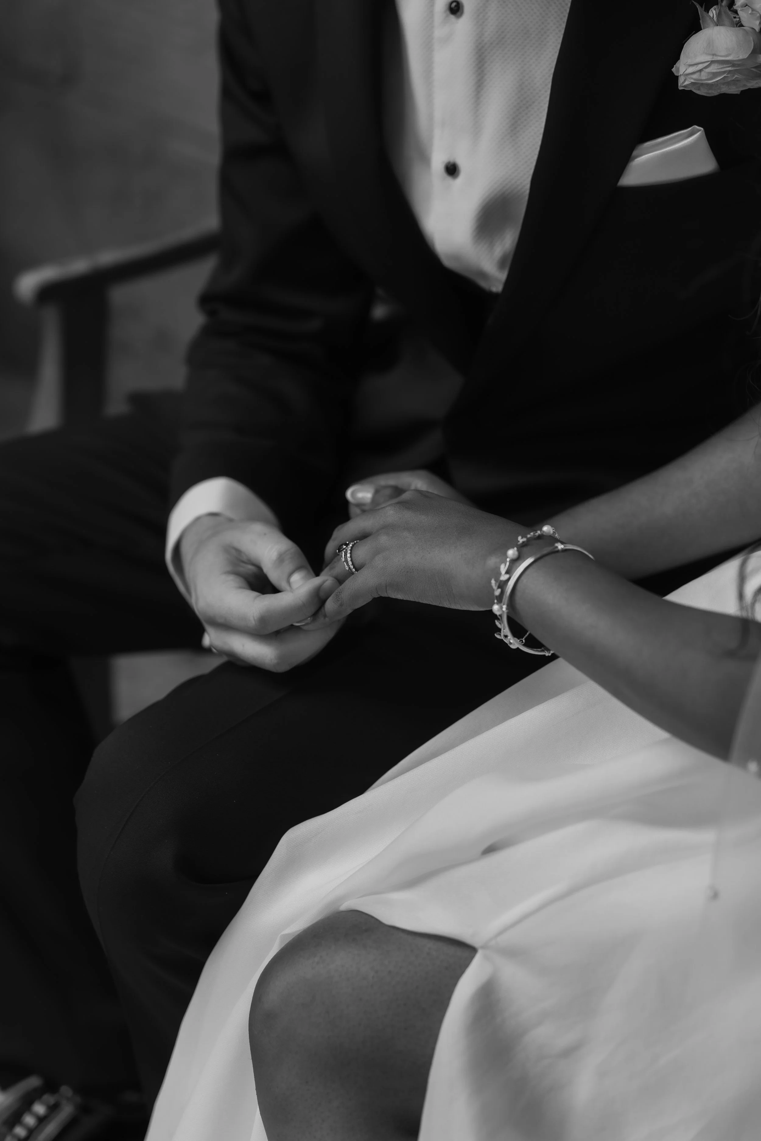 A black and white photo of a couple holding hands, with the man wearing a suit and the woman wearing a dress, wearing bracelets and a ring.