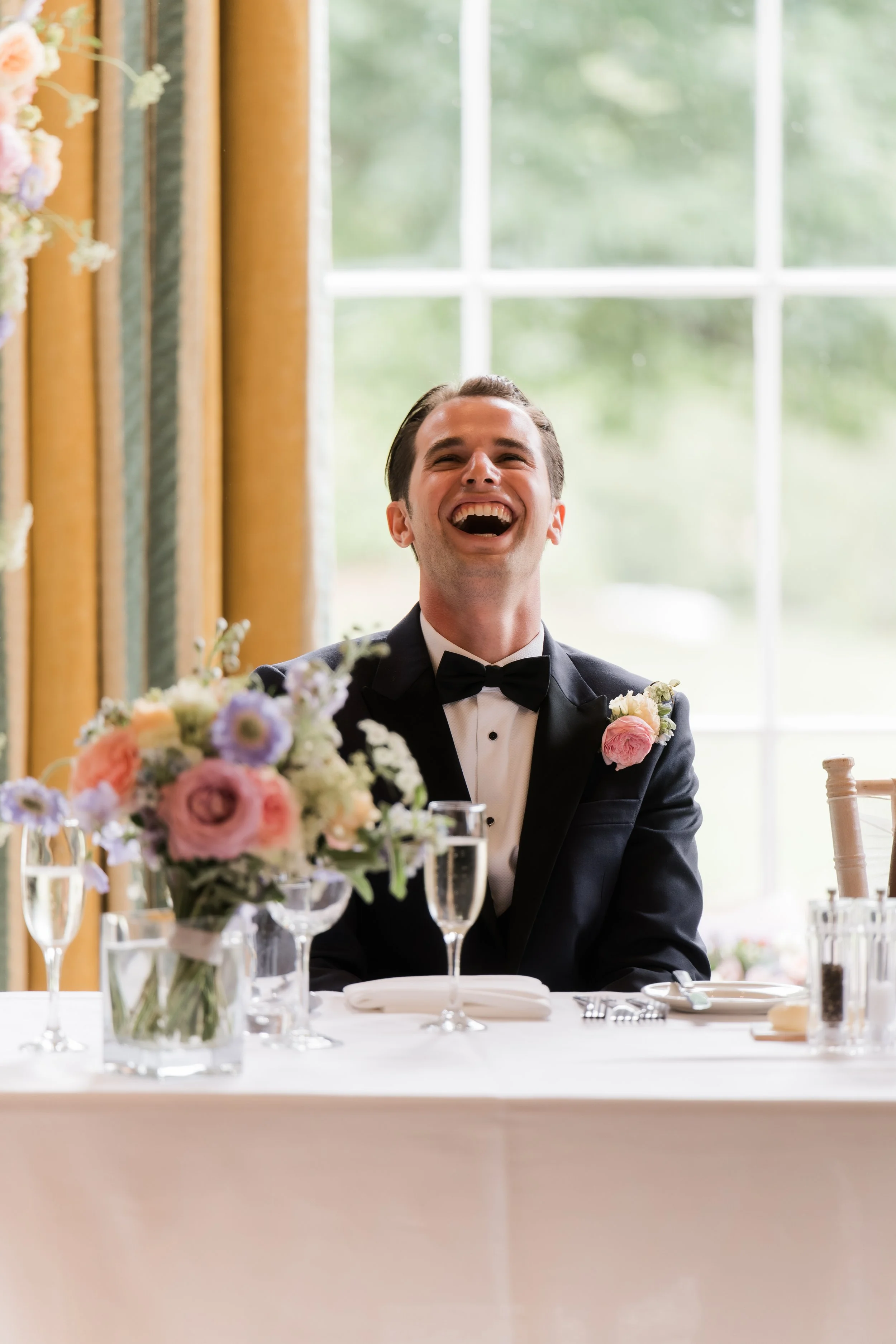 A man in a tuxedo with a bow tie, laughing joyfully while sitting at a decorated table with a floral centerpiece, in a well-lit room with large windows.