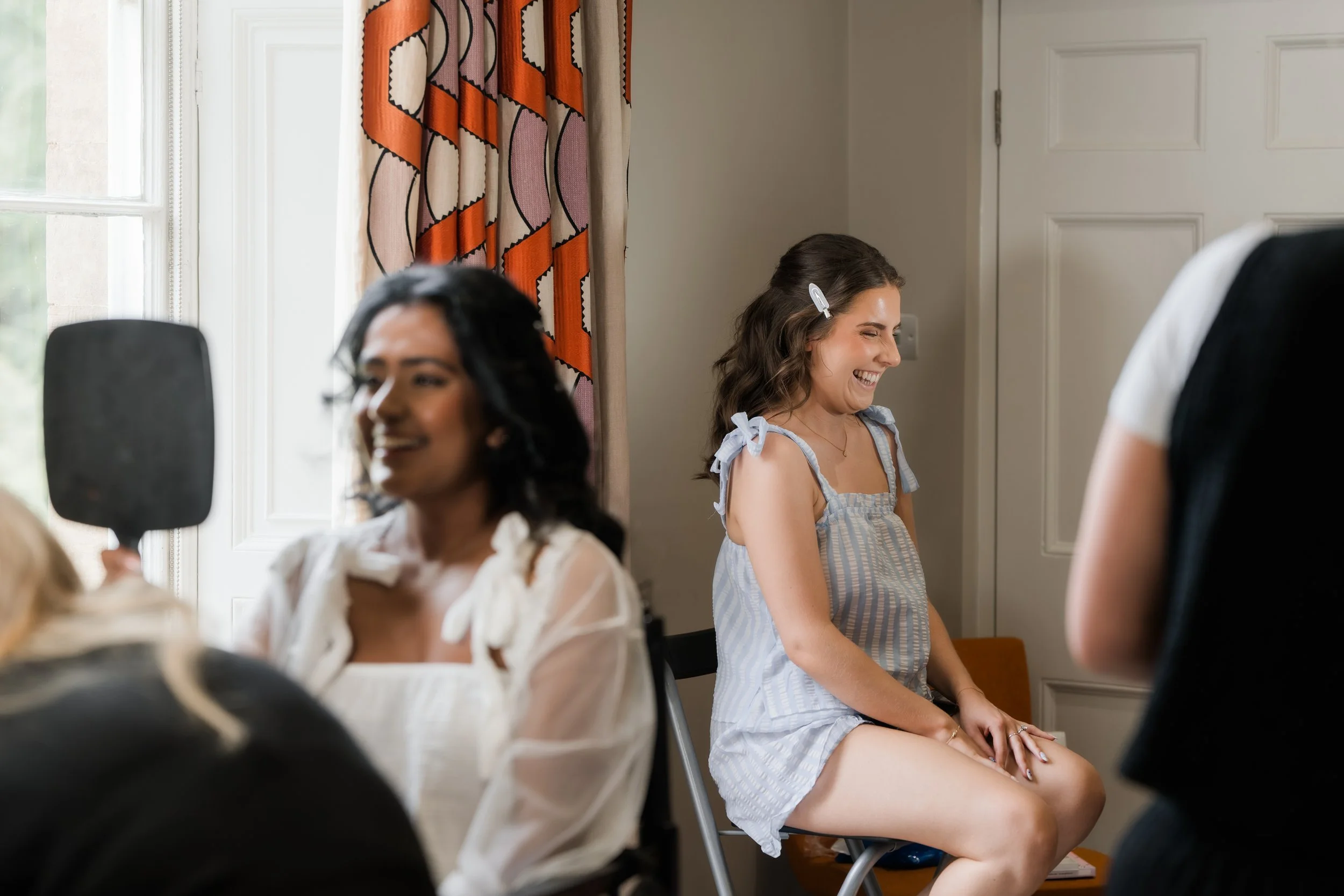 Three women sitting indoors, one with a clip in her hair, all smiling and talking, with a window and colorful curtains in the background.