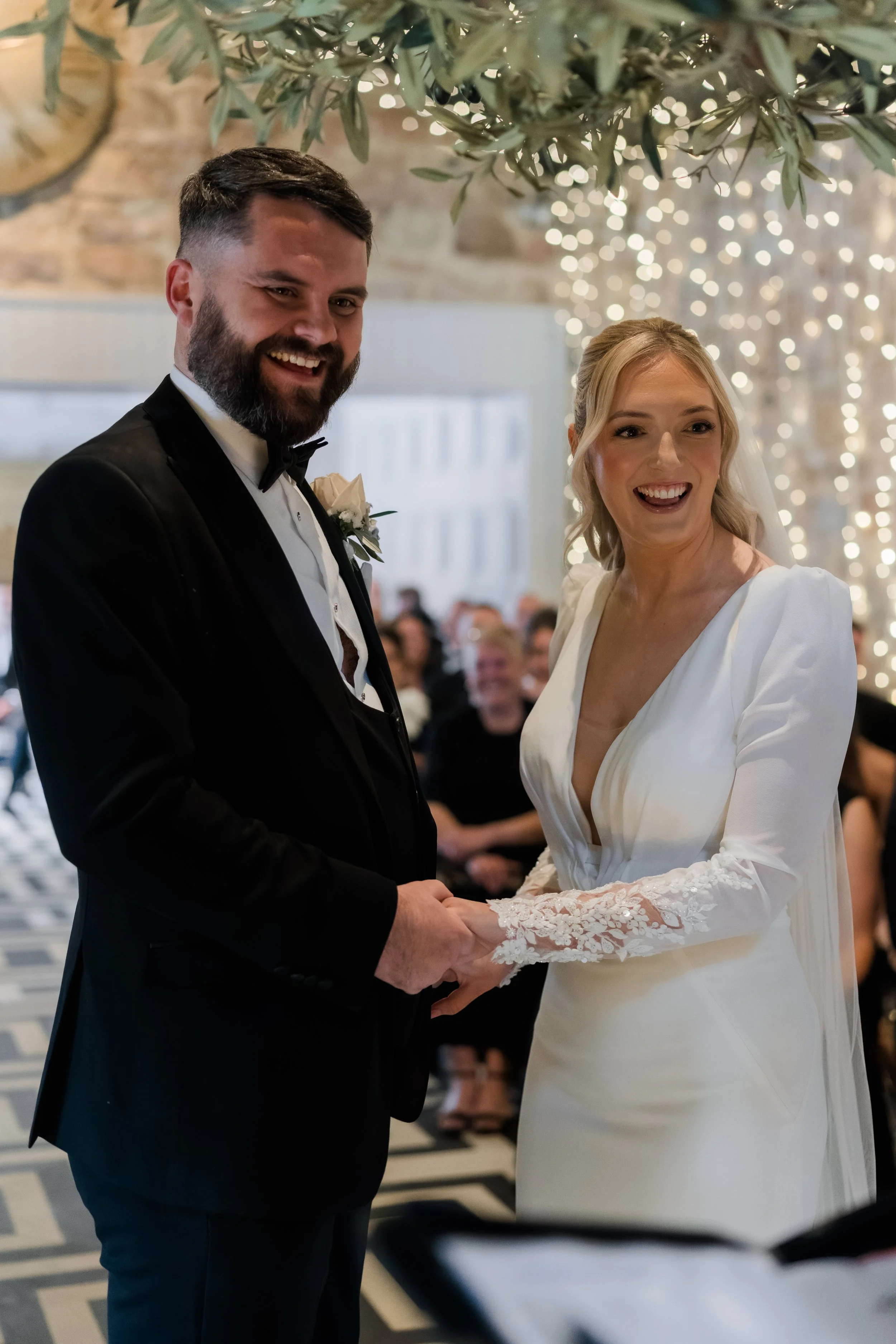 A bride and groom holding hands during their wedding ceremony, smiling happily. The bride wears a white dress with lace sleeves, and the groom wears a black tuxedo with a bow tie.