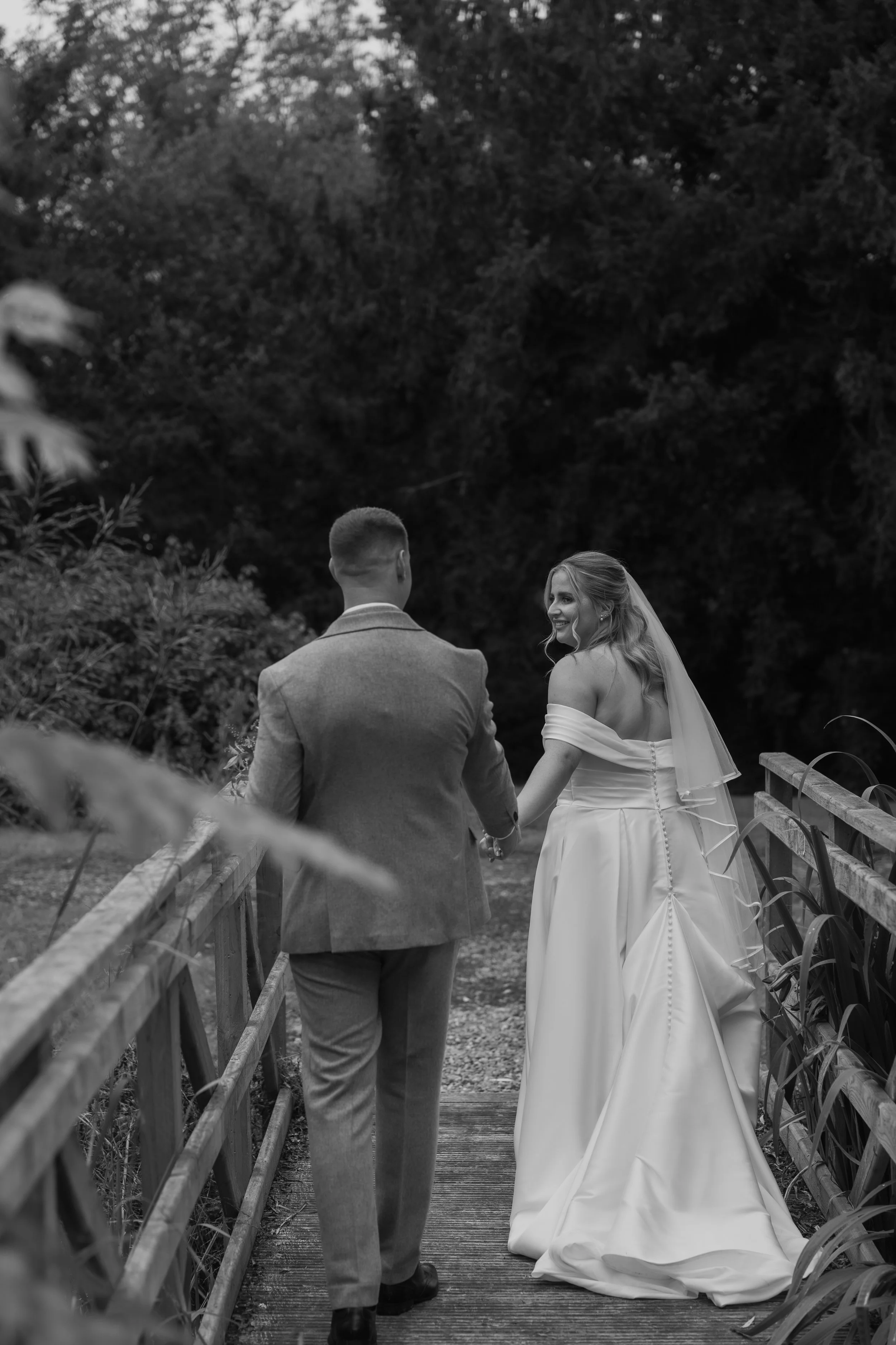 A black and white photo of a bride and groom holding hands while walking on a wooden bridge outdoors, surrounded by trees and foliage.