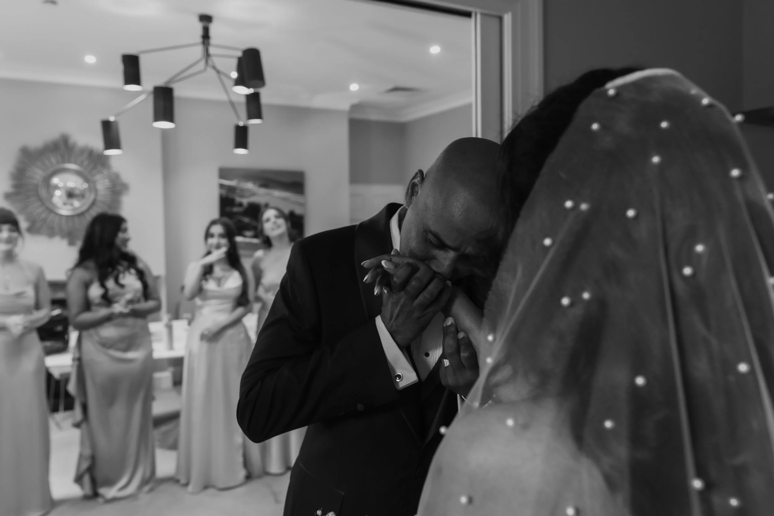 Man in a tuxedo kissing a woman in a wedding dress at a wedding reception with bridesmaids in the background.