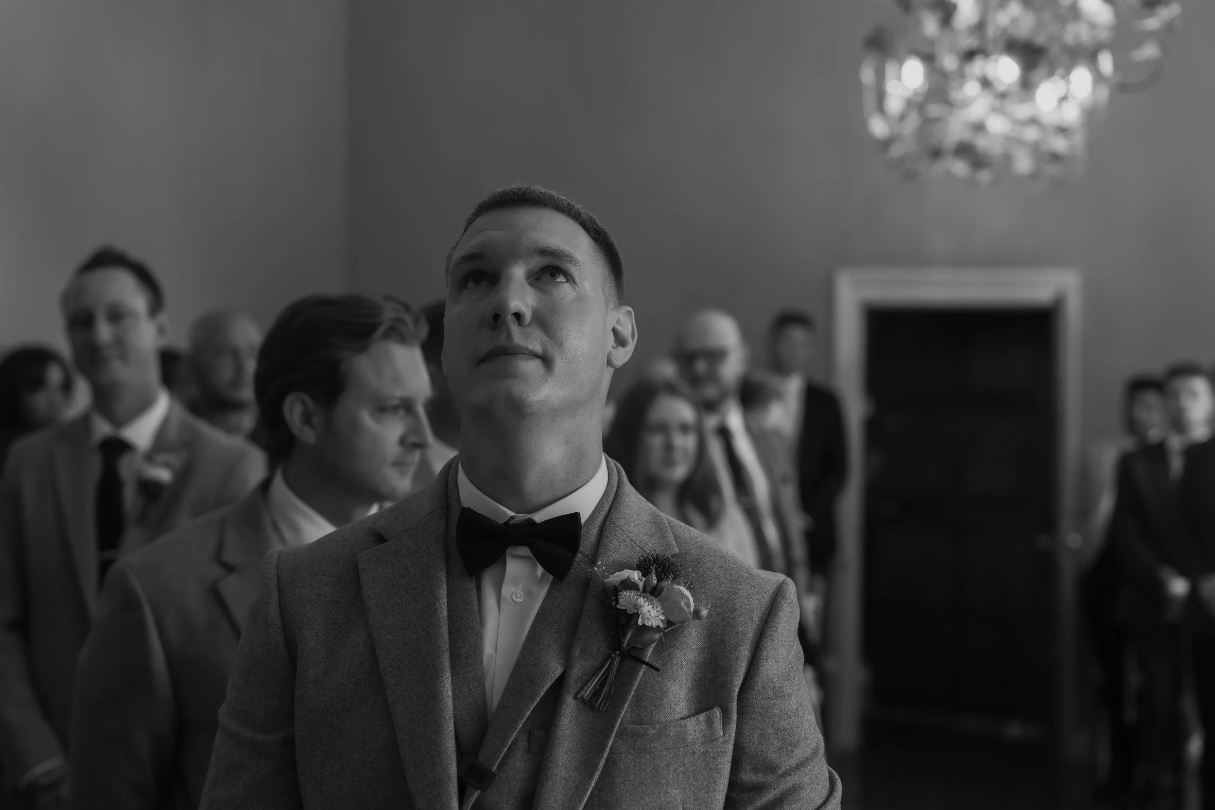 Black and white photo of a groom in a suit with a flower boutonniere, looking upwards during a wedding ceremony, with guests in the background.