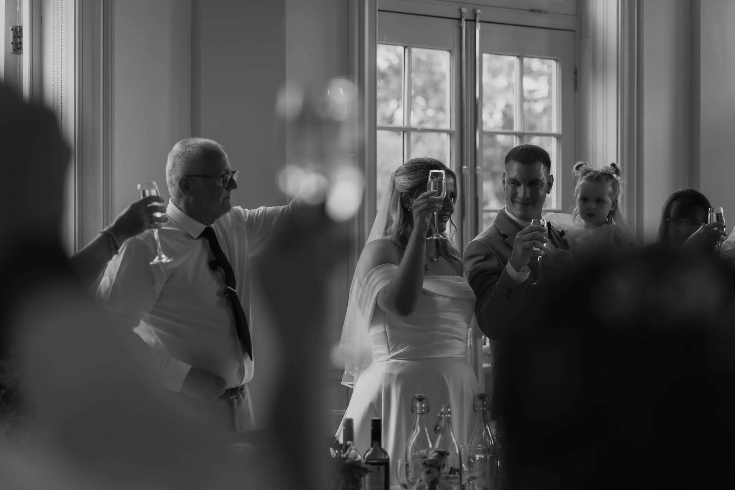 Black and white champagne toast to the bride and groom shot through the guests raised arms. Hodsock Priory.  Nottinghamshire.