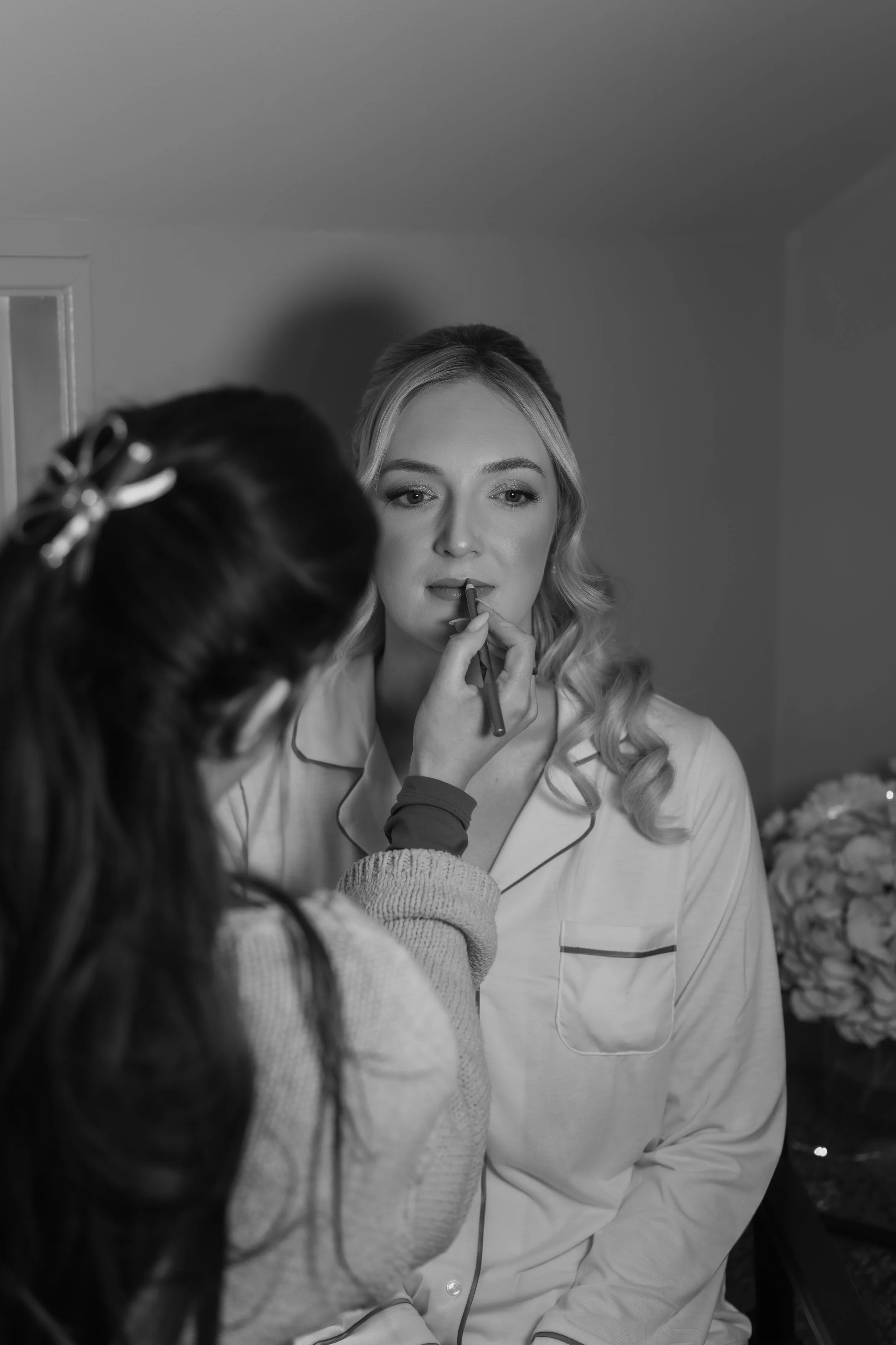A woman with light-colored, wavy hair wearing pajamas is having her makeup done by a makeup artist with dark hair and a hair clip.