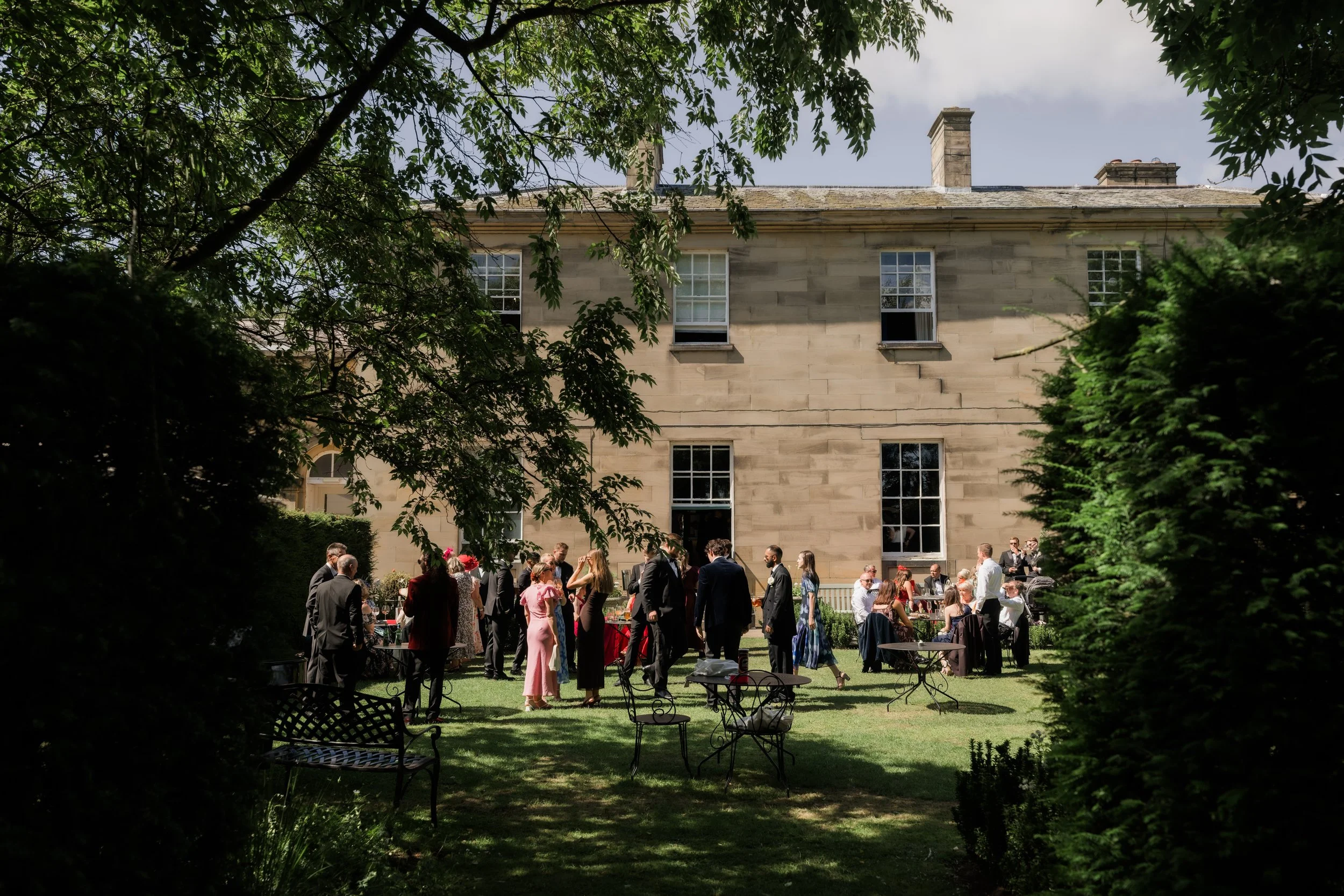 A wedding reception in a lush outdoor garden with guests mingling, some standing and some seated, near a historic stone building with multiple windows and three chimneys, framed by trees and greenery.