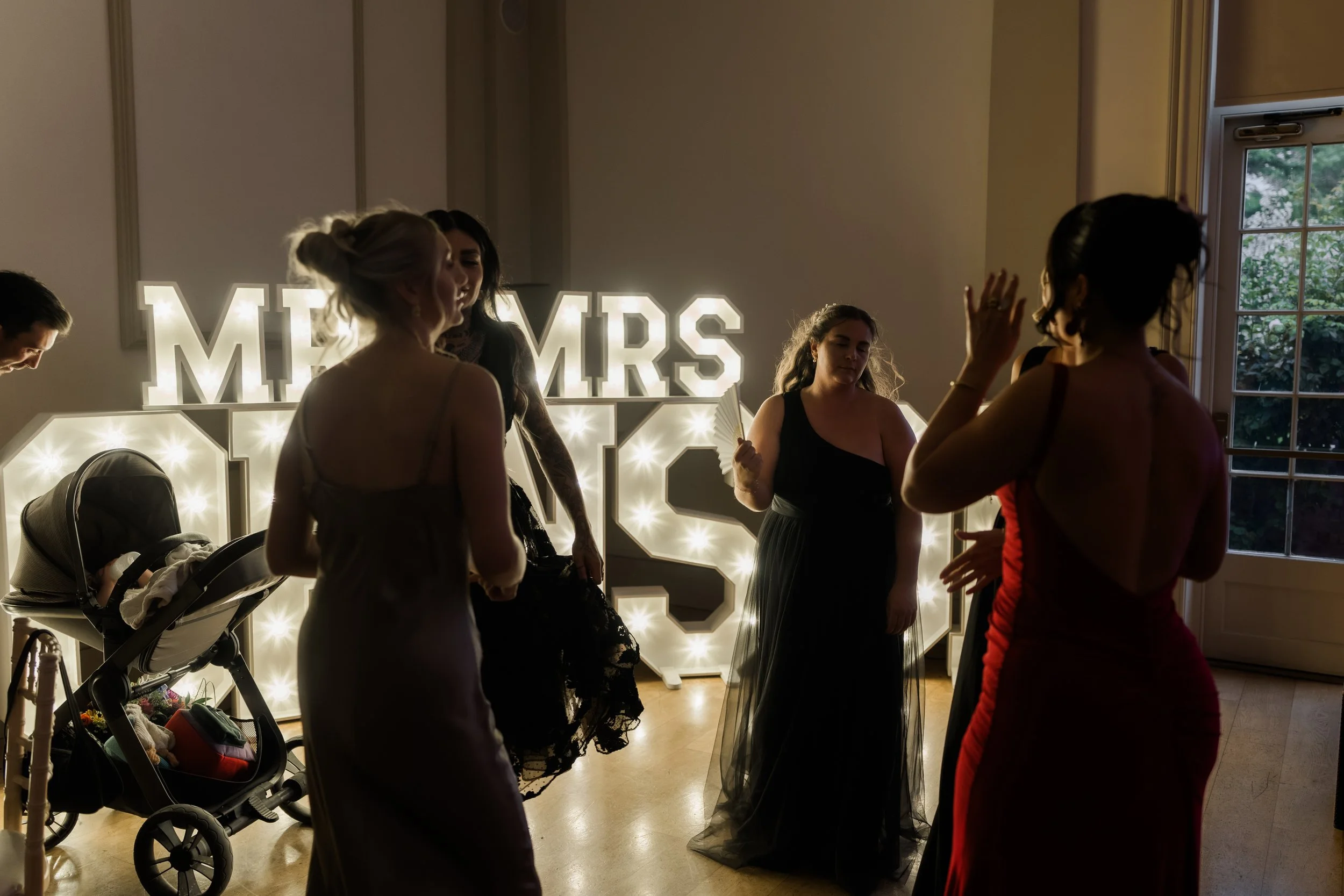 Wedding reception with guests dancing, a lit 'MRS' sign, and a woman holding a fan.