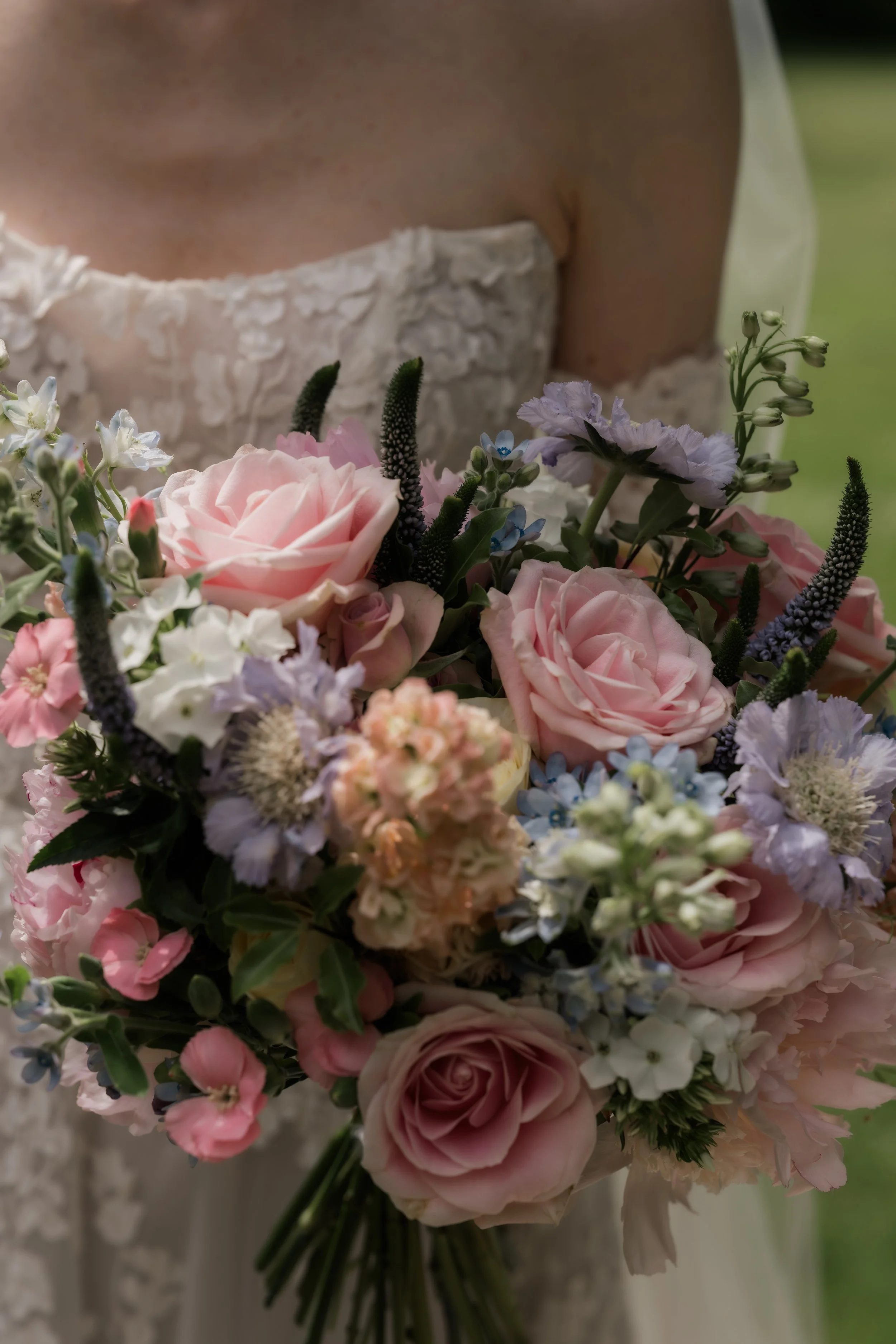 A close-up of a bridal bouquet with pink roses, white and light purple flowers, green leaves, and other small flowers.