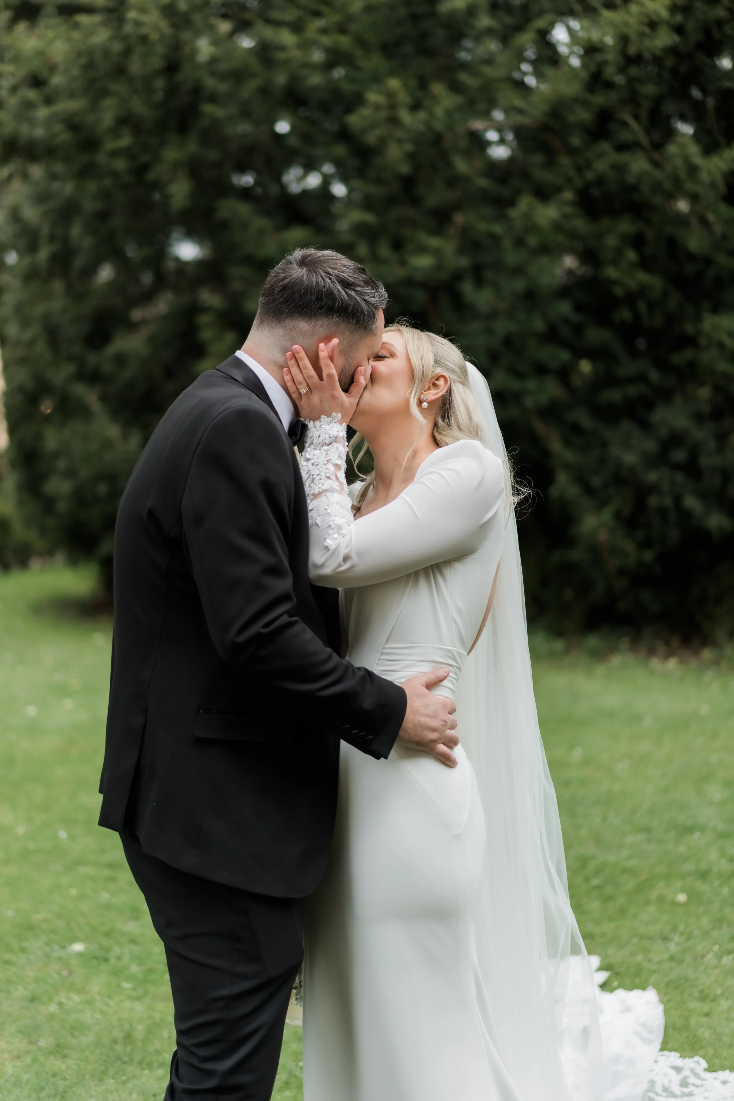 A bride and groom kiss outdoors on grass with green trees in the background.