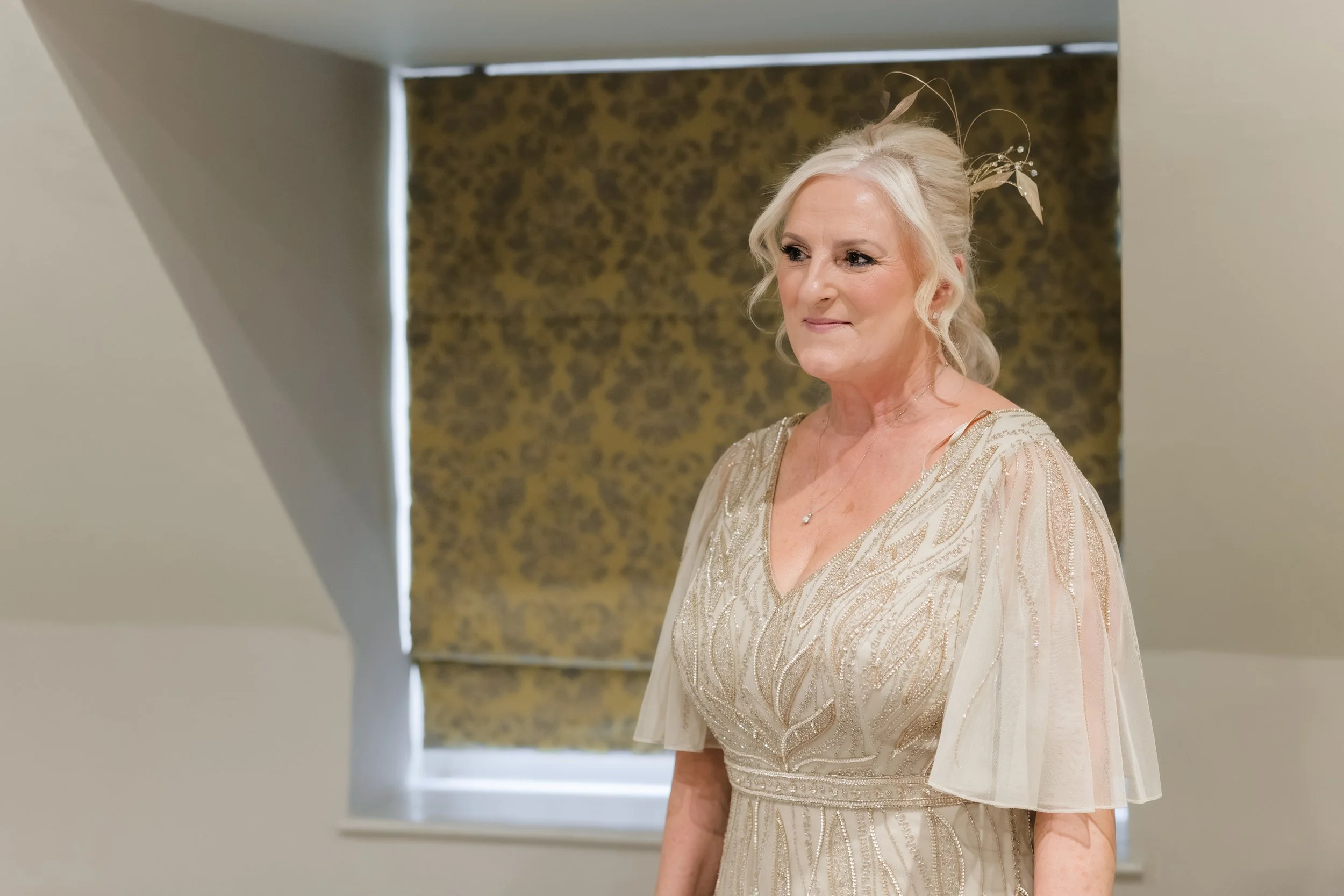 An elderly woman with blonde hair styled in curls, wearing a light-colored, ornate dress with embroidered details, stands near a window with a patterned curtain in the background.