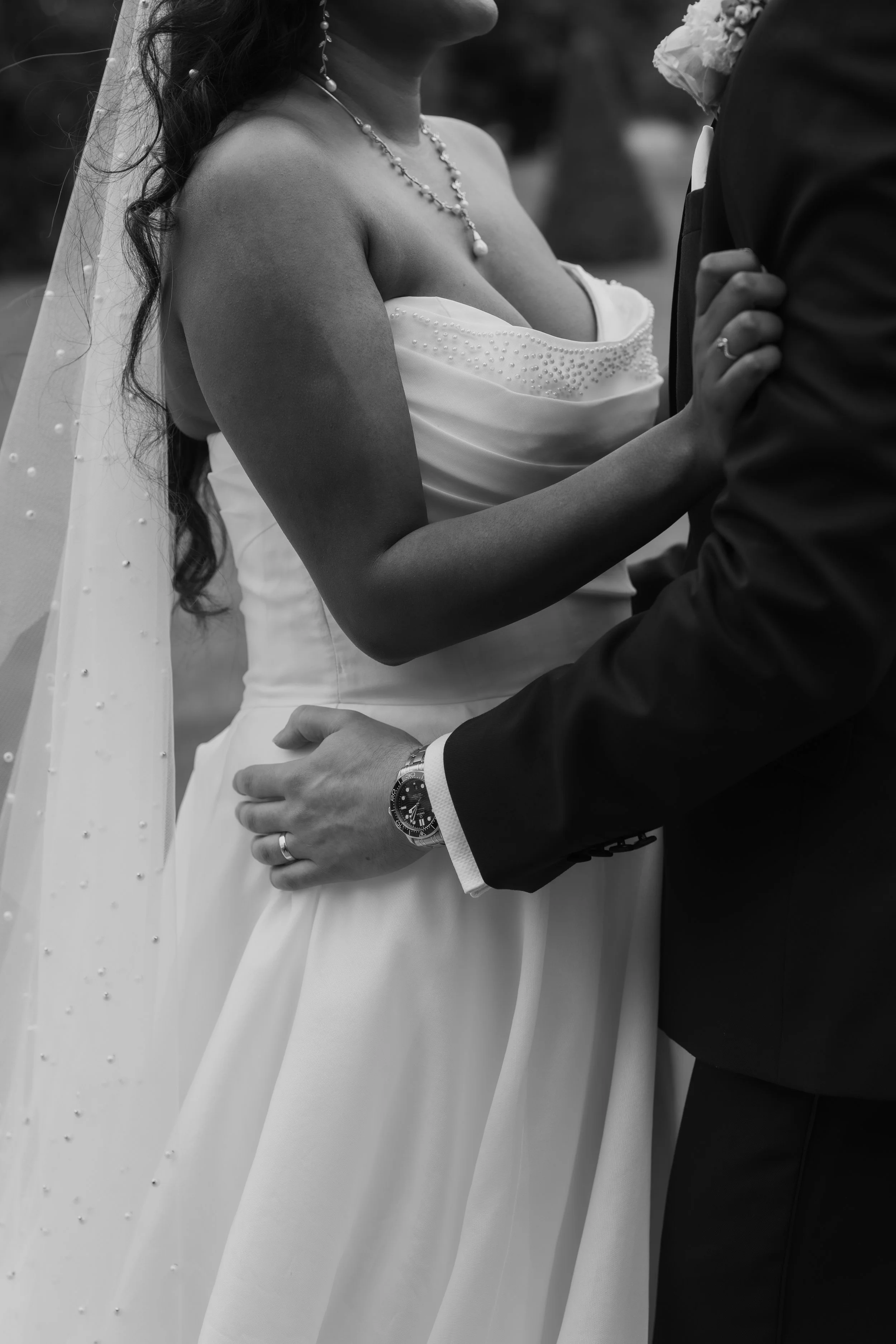 A bride and groom standing close together holding hands during their wedding, with the bride wearing a strapless wedding dress, veil, and jewelry, and the groom wearing a black suit and watch.