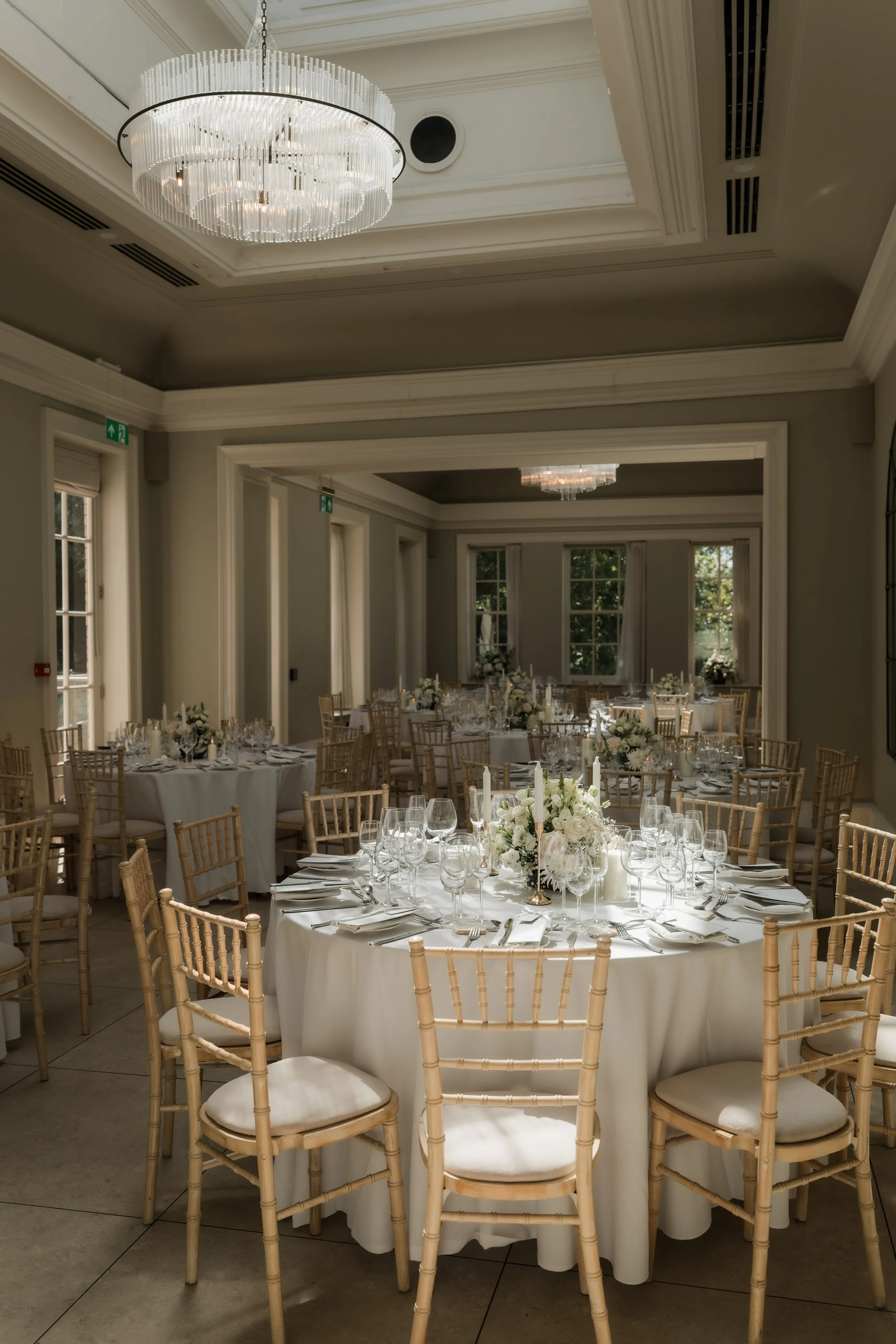 Elegant banquet hall decorated for a wedding reception with round tables, white tablecloths, floral centerpieces, candles, and gold chairs. Crystal chandeliers hang from the ceiling, and large windows allow natural light into the room.