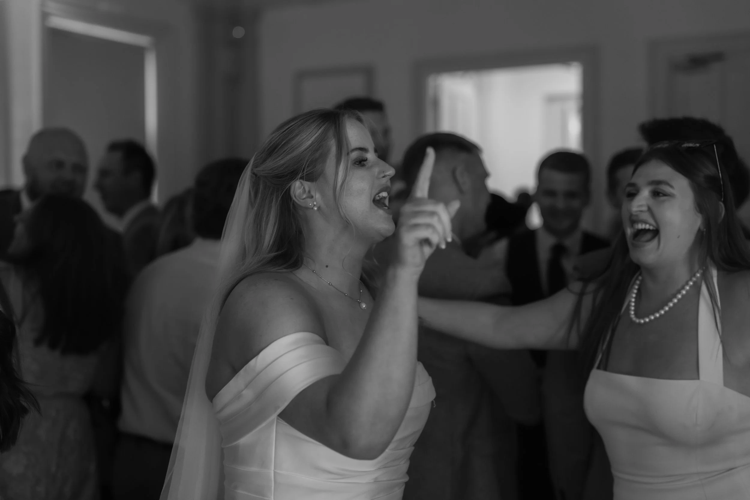 A woman in a wedding dress dancing and singling with another woman at a wedding reception, surrounded by guests in a decorated room.