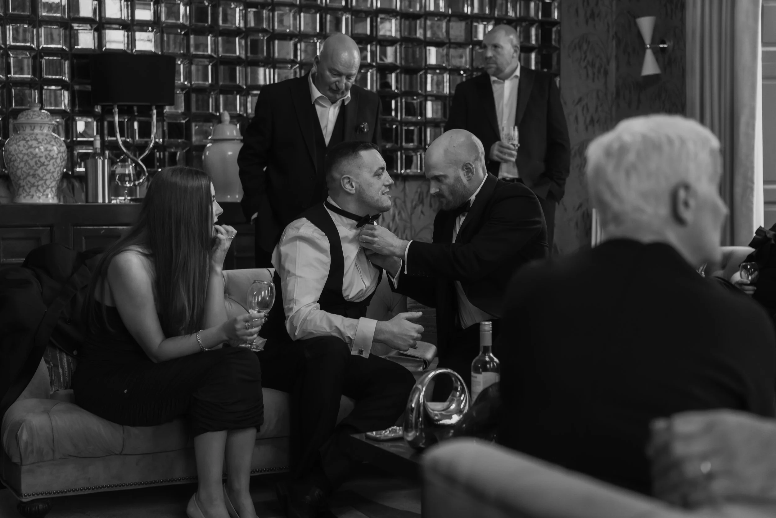 A group of people at a formal gathering or party, with a person adjusting another person's bowtie, while others sit and stand holding drinks, in a room decorated with vases, bookshelves, and curtains.