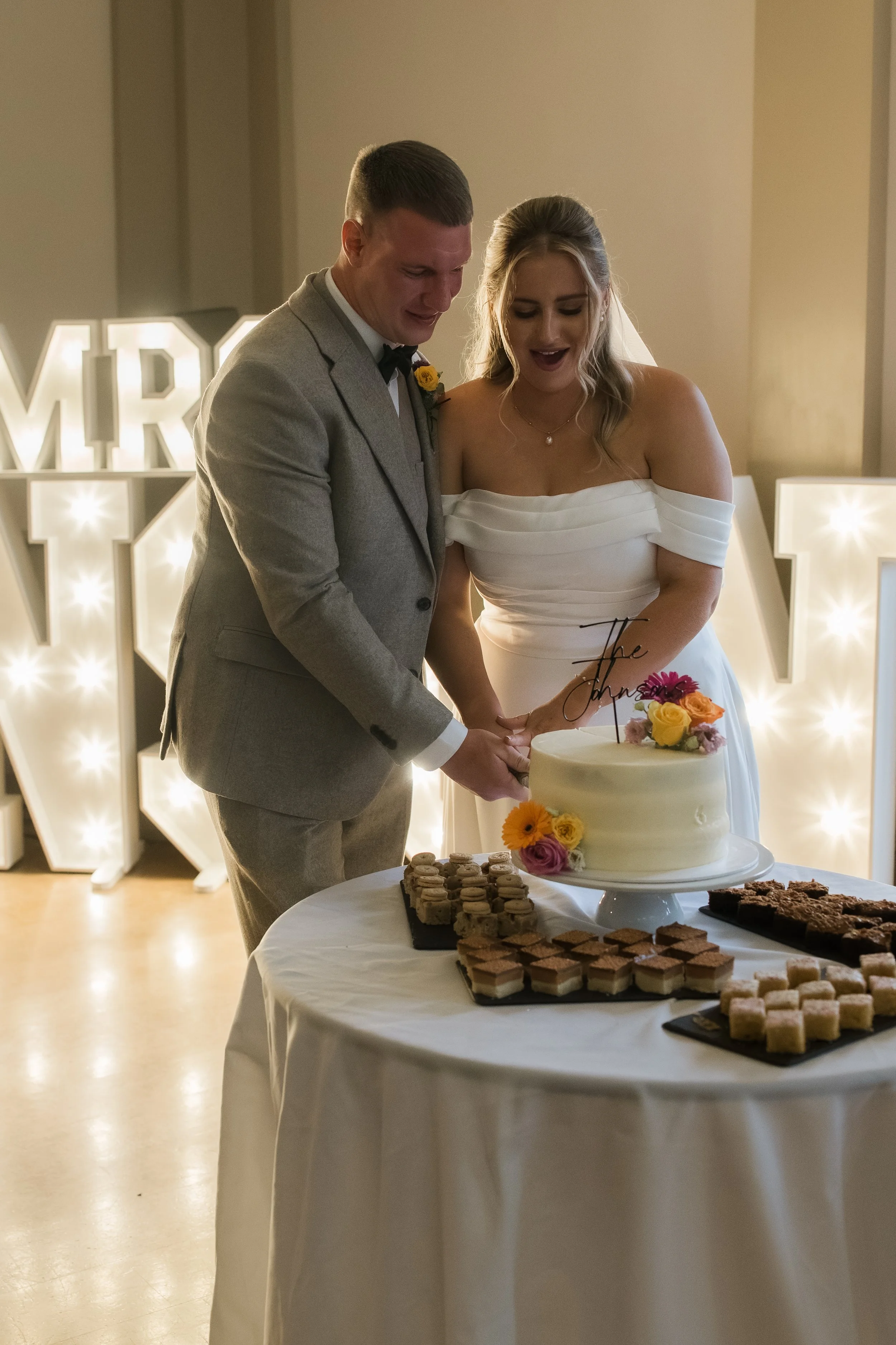 A newly married couple is cutting a wedding cake at their reception. The groom is dressed in a gray suit with a bow tie, and the bride is in a white off-the-shoulder wedding gown. They are smiling and holding a cake knife together. There are various 