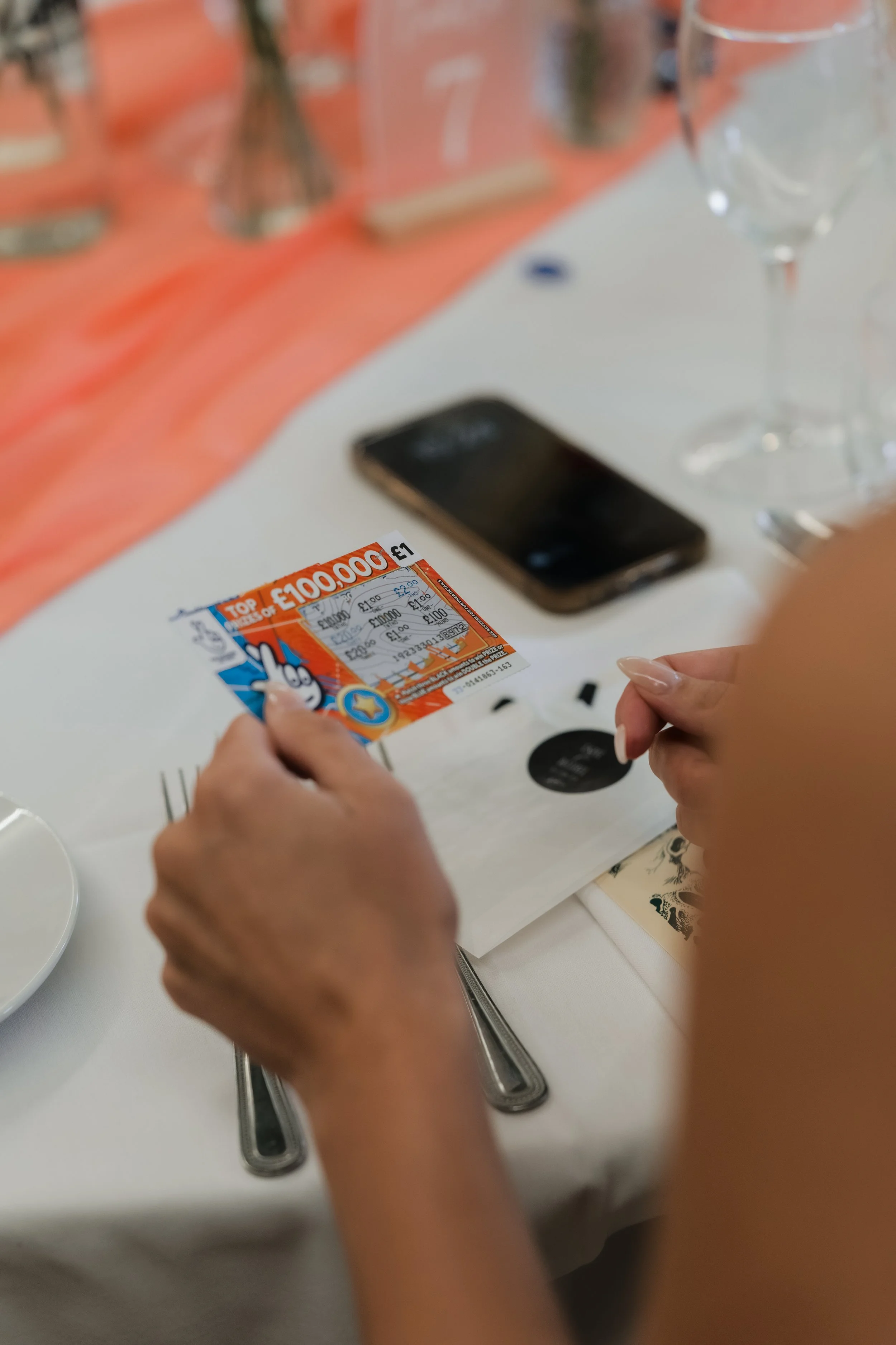 Person holding a scratch-off lottery ticket at a table set for a meal with wine glasses, chopsticks, and a smartphone.