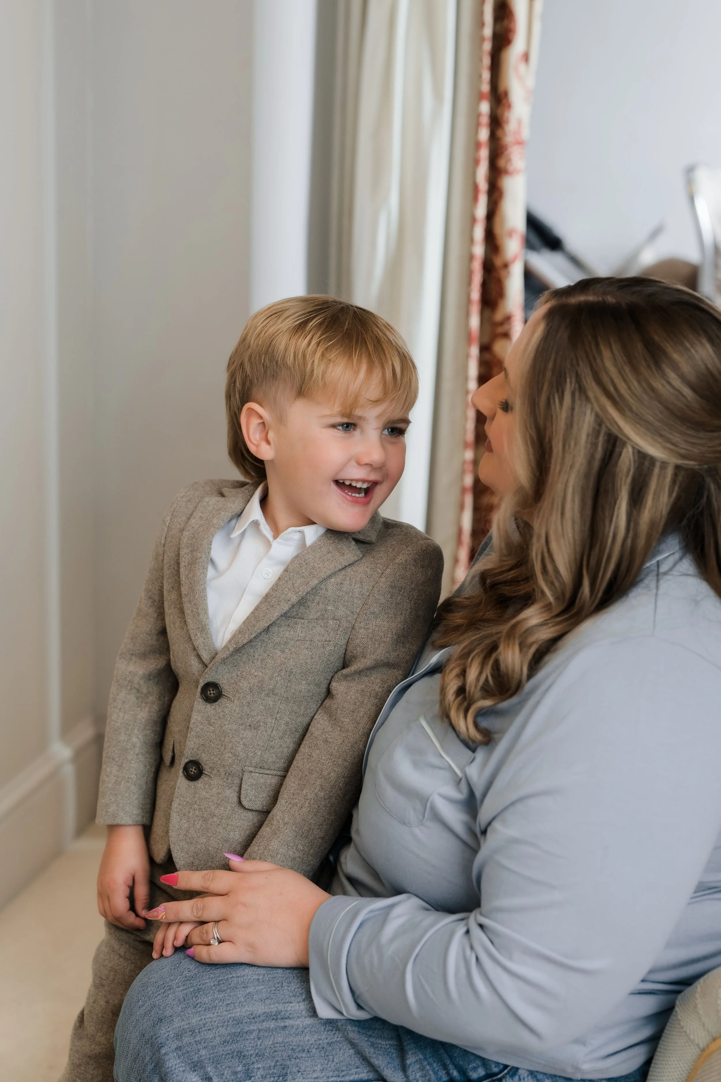 A young boy in a beige suit smiling and talking to a woman sitting on a chair, with curtains in the background.