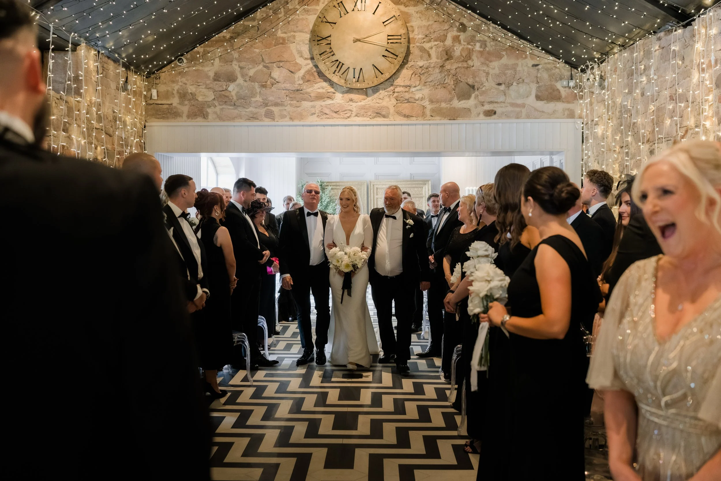 A wedding ceremony with the bride in a white dress holding a bouquet, escorting by two men, one larger man with sunglasses and the other with a beard. The guests are dressed in black and are standing on either side of the aisle, some holding white fl