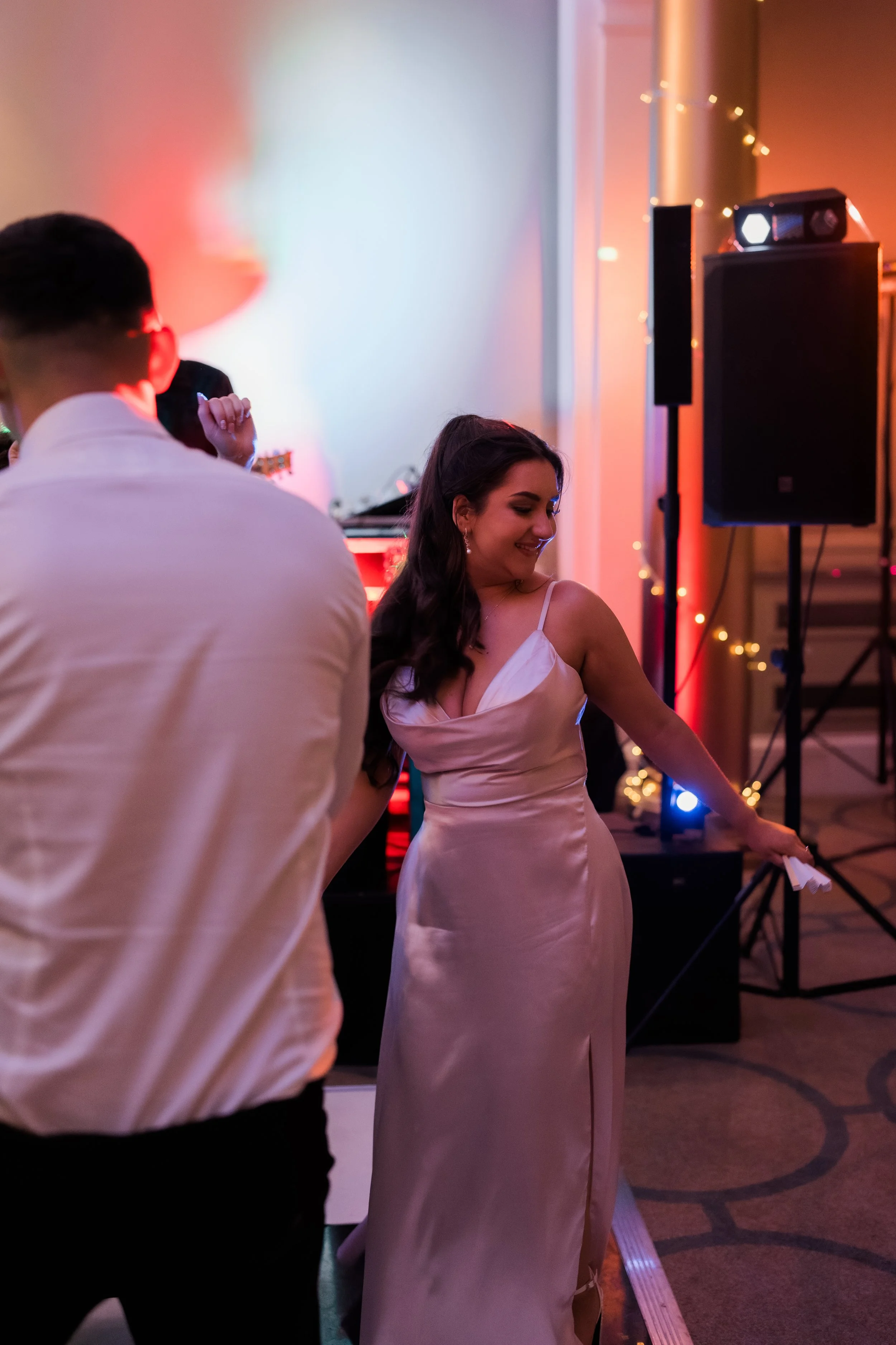 A woman dancing at a party or celebration in a satin dress with spaghetti straps, smiling and holding a small object.