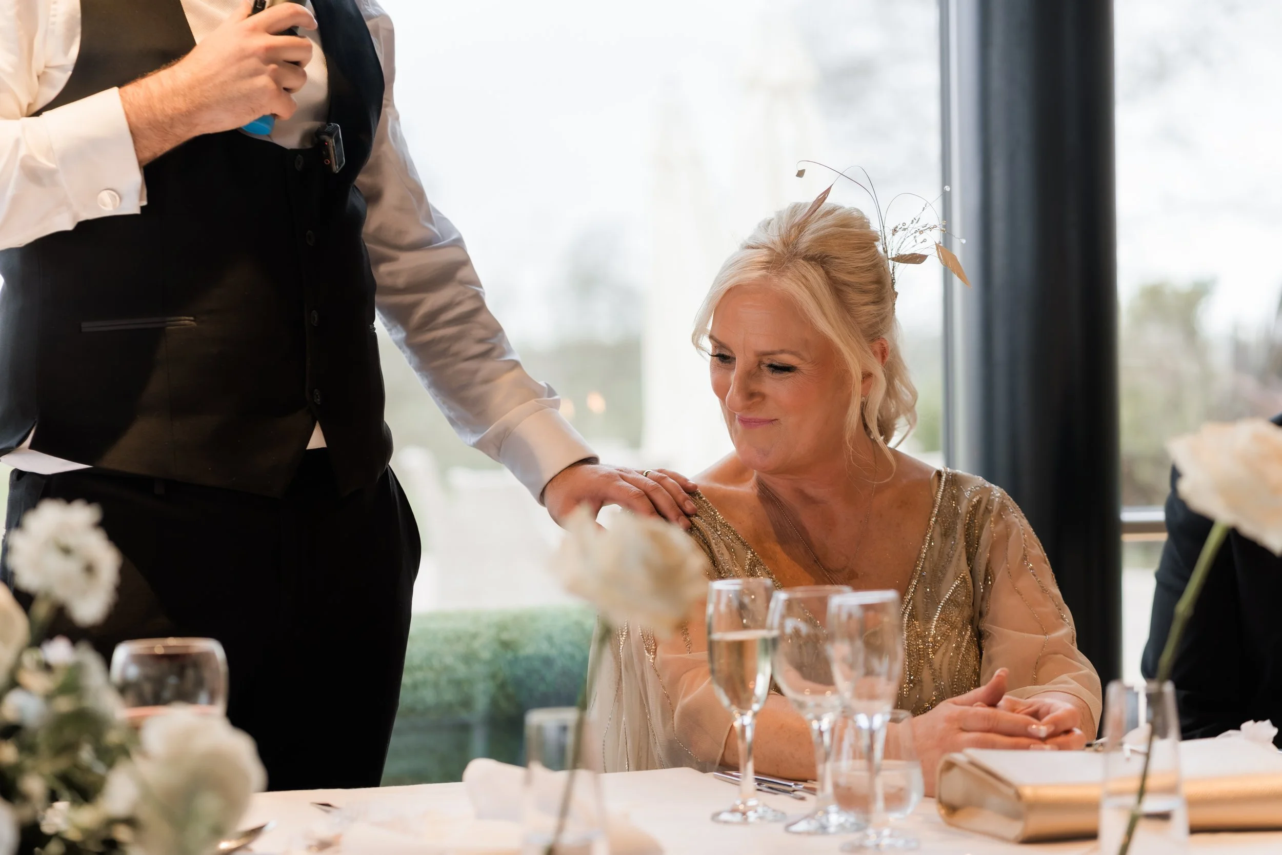 A woman with blonde hair and a golden dress sitting at a table while a waiter touches her shoulder during a celebration.