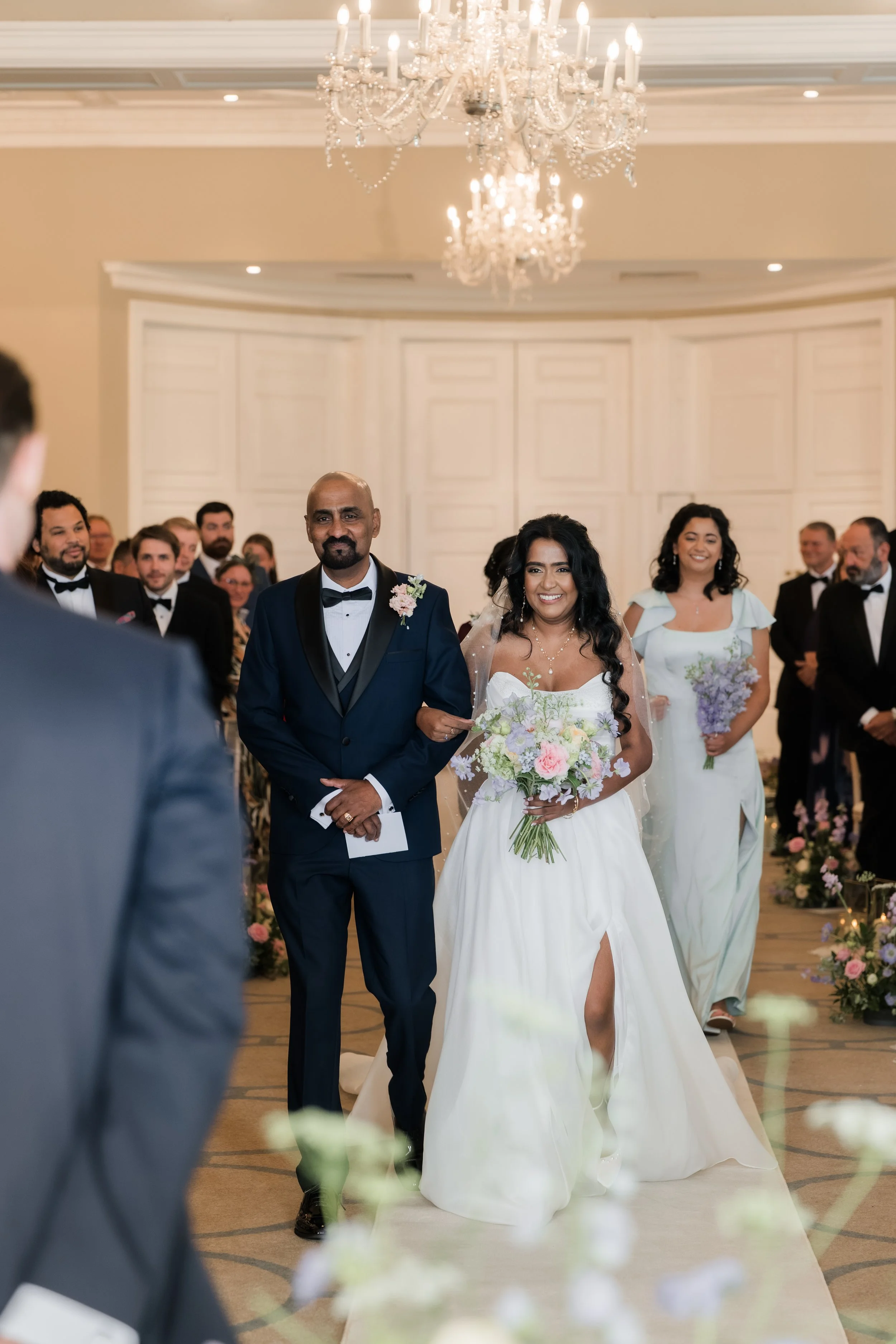A bride walking down the aisle with a man in a tuxedo at her side, surrounded by wedding guests in formal attire, in an elegant decorated venue with chandeliers.