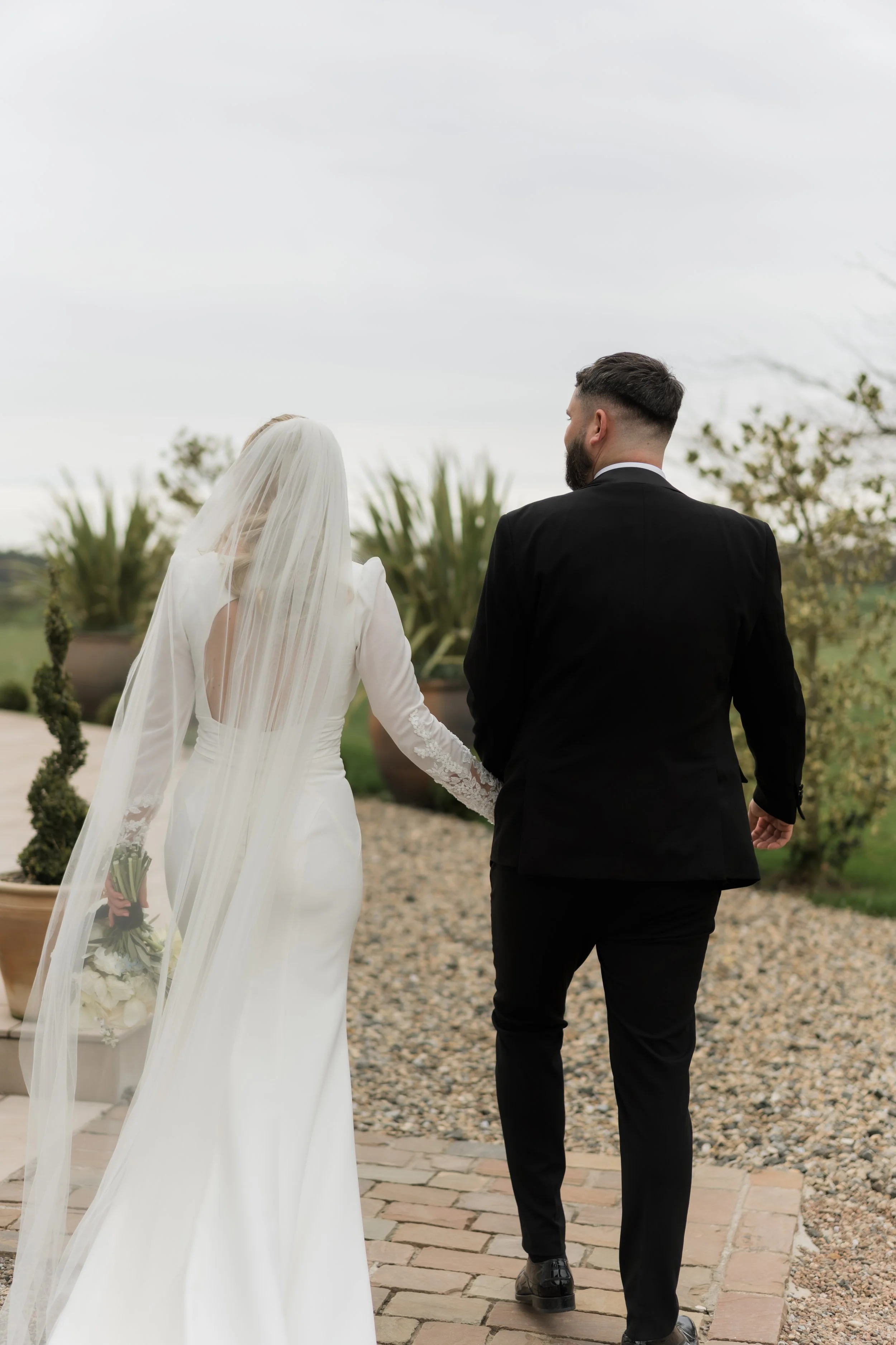 A bride and groom walking hand in hand outdoors during their wedding, with the bride in a white dress and veil holding a bouquet, and the groom in a black suit, on a brick pathway surrounded by plants.