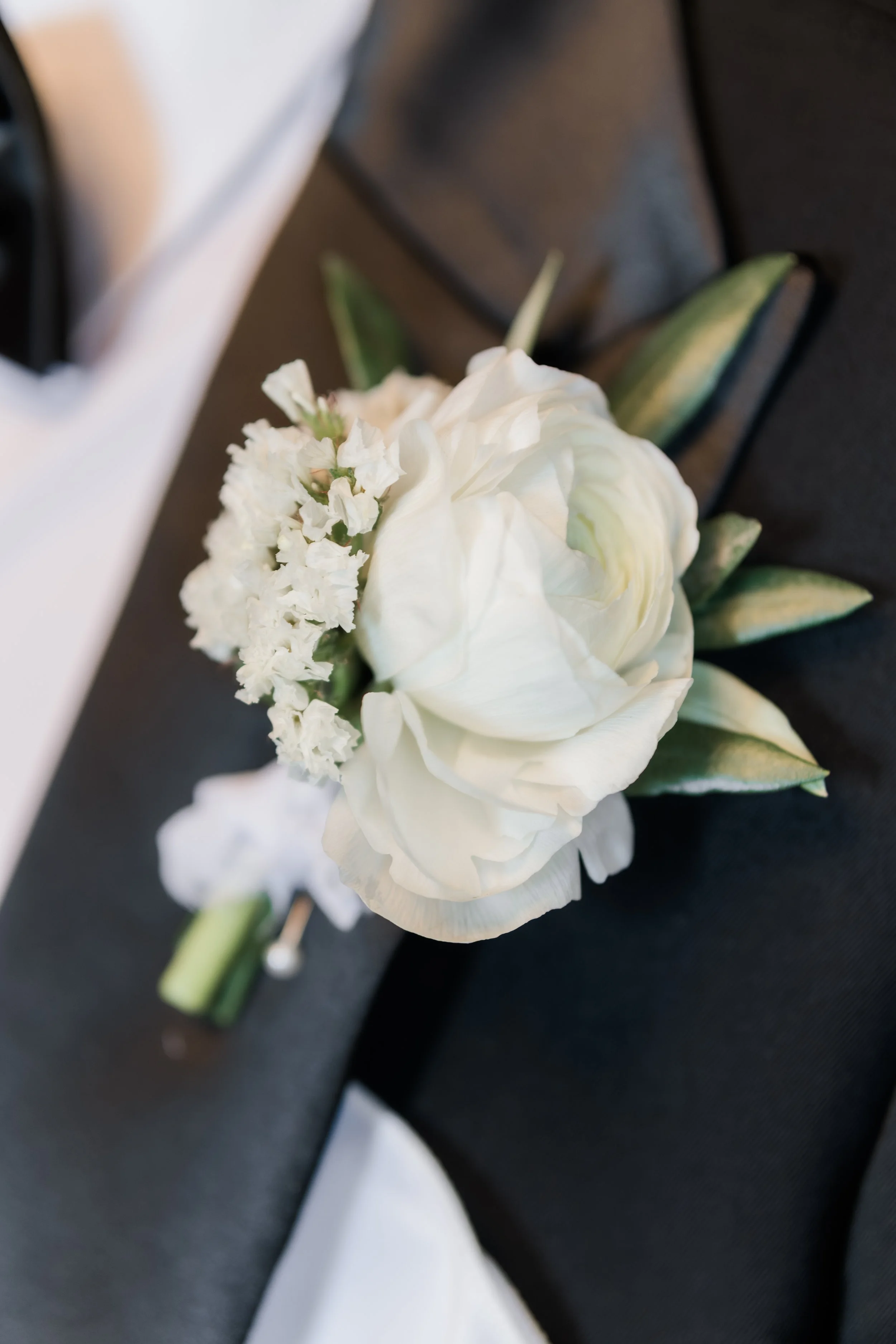 Close-up of a white boutonniere with a white flower and green leaves on a black surface.