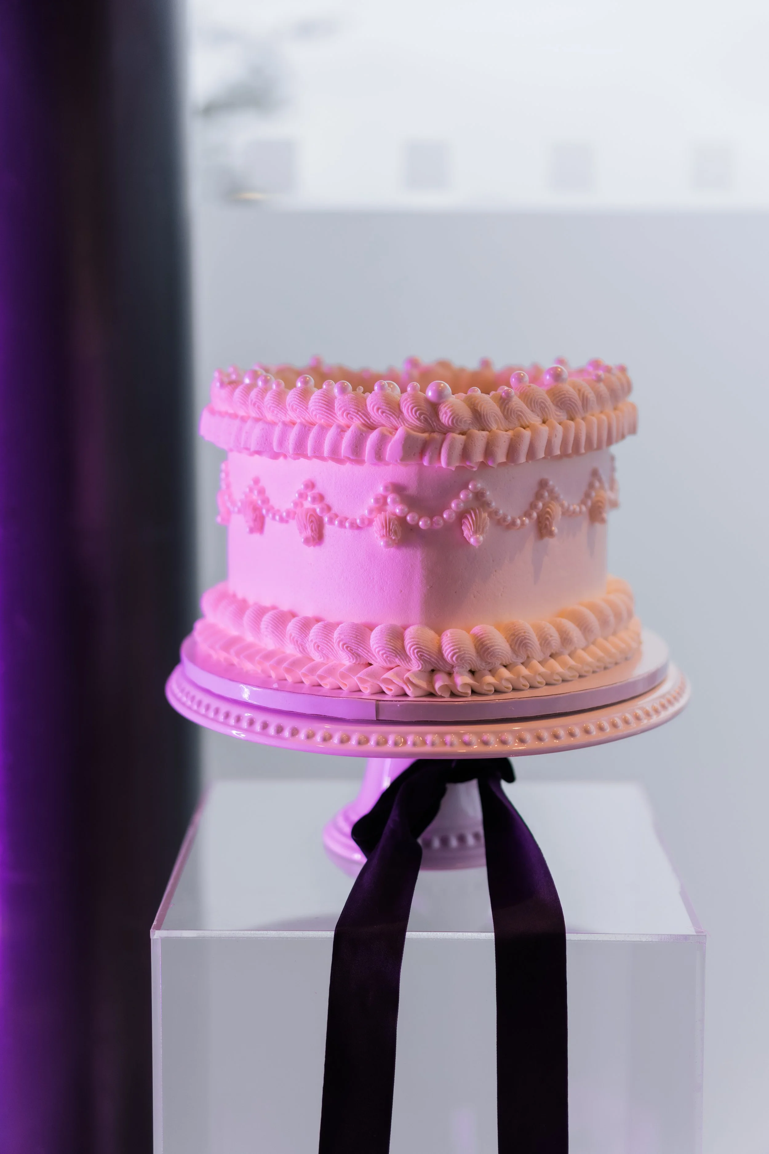 A pink, decorated birthday cake on a stand with a black ribbon, displayed on a white table.