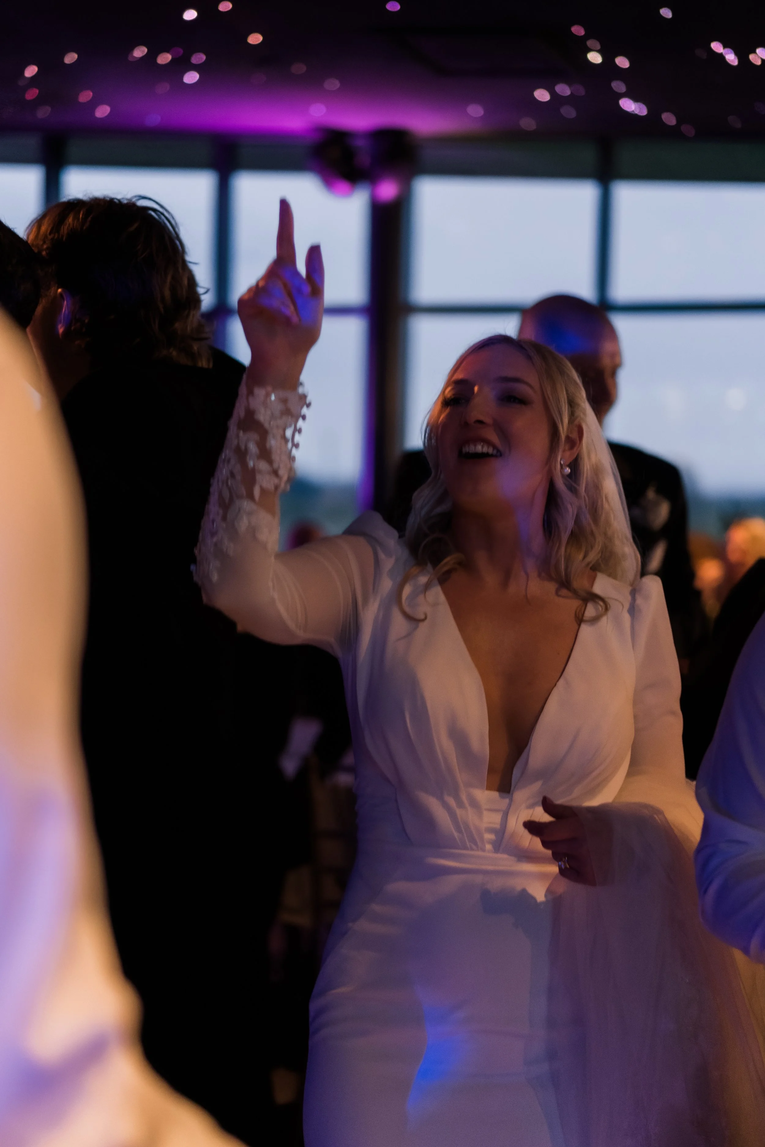 A woman in a white dress dancing and smiling at a party, with other people around her in a dimly lit room with purple lighting and large windows.
