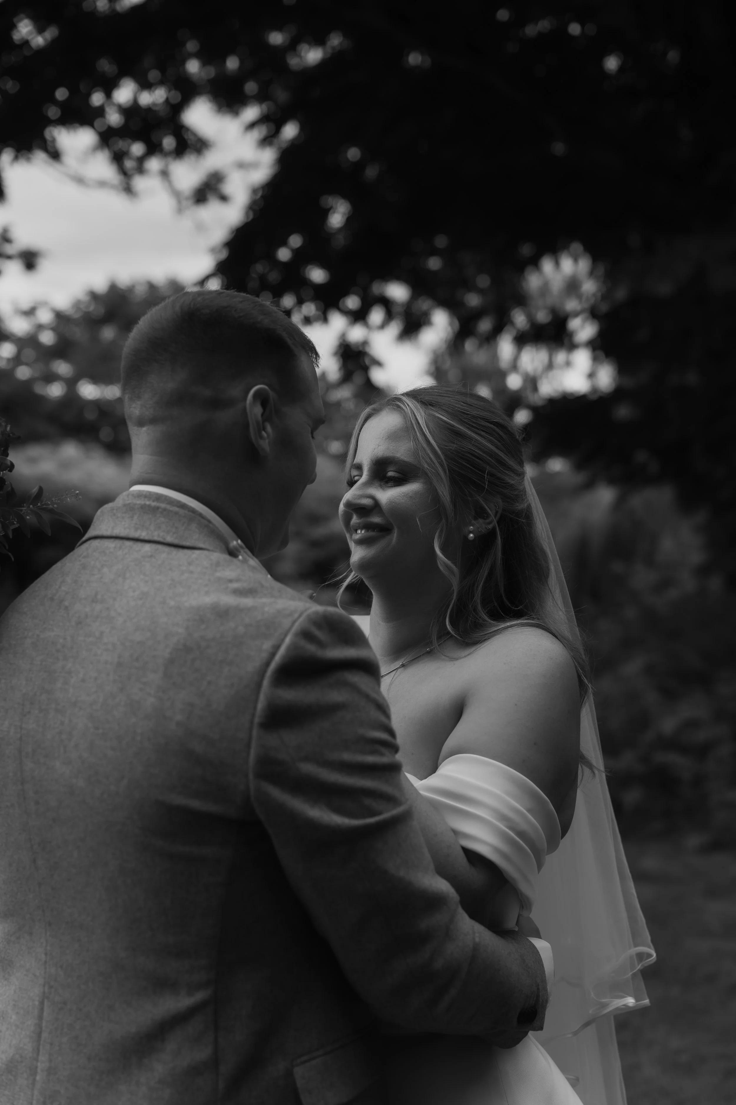 A black and white photo of a bride and groom embracing outdoors, smiling at each other with trees in the background.