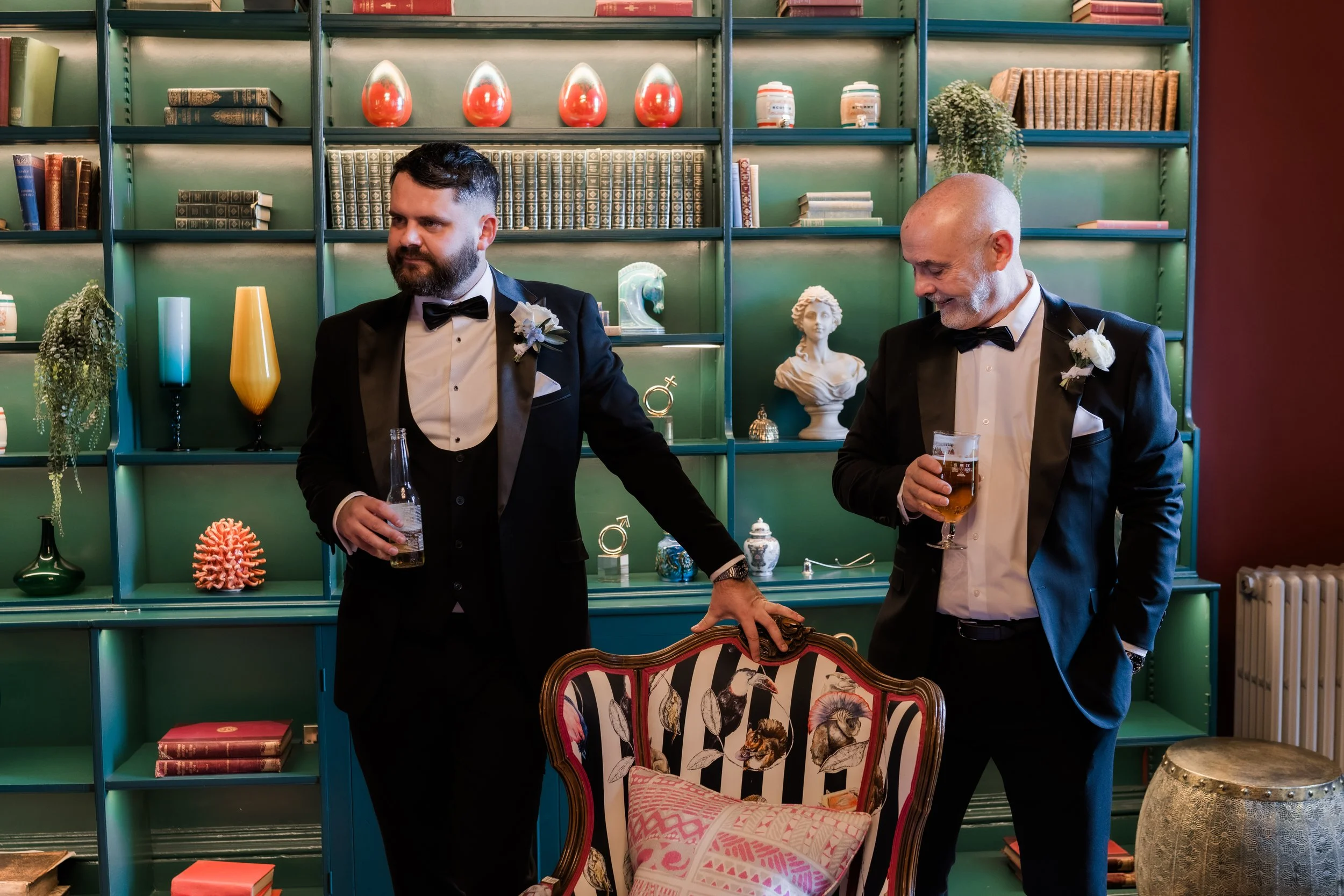 Two men in tuxedos with boutonnières standing indoors, holding drinks, one with a beard and the other bald with a gray beard, against a green bookshelf filled with decorative objects, books, and colorful vases.