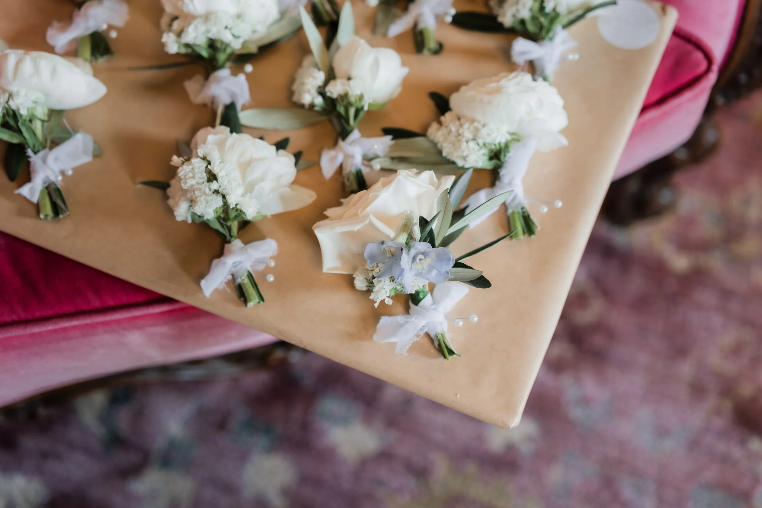 A table with cream-colored floral boutonnieres and corsages, including white roses, baby’s breath, and greenery, attached with white ribbons and pins, on brown wrapping paper.