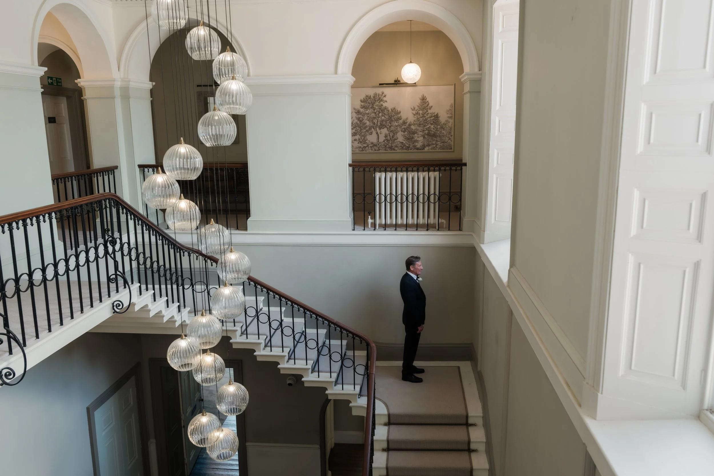 Interior view of a grand staircase with modern hanging lights and white walls. A man in a tuxedo stands on the lower level near a radiator, looking upward.