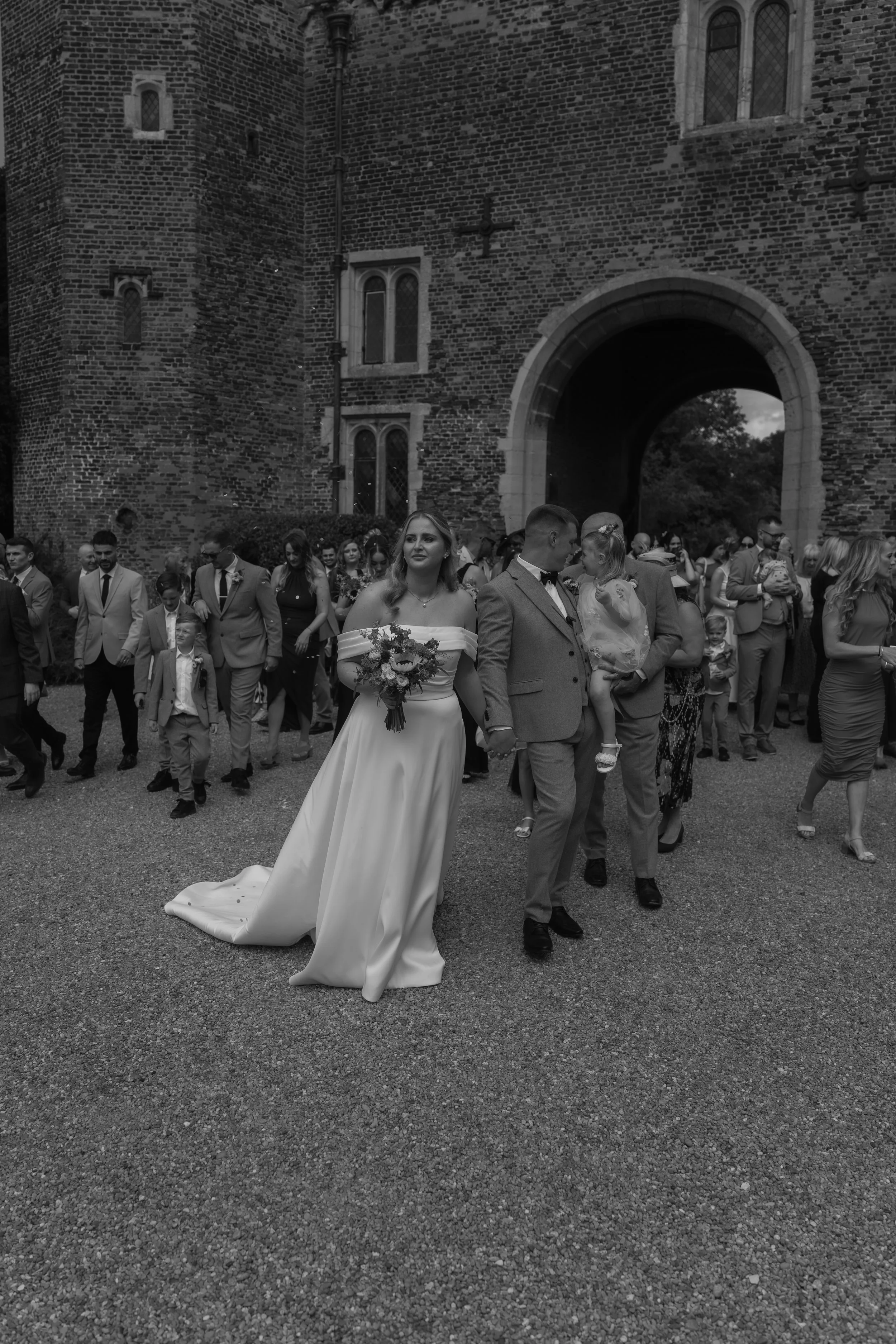 A wedding procession outside a historic brick church with a large arched entrance, including a bride in a white gown holding a bouquet and a groom in a gray suit, surrounded by guests.