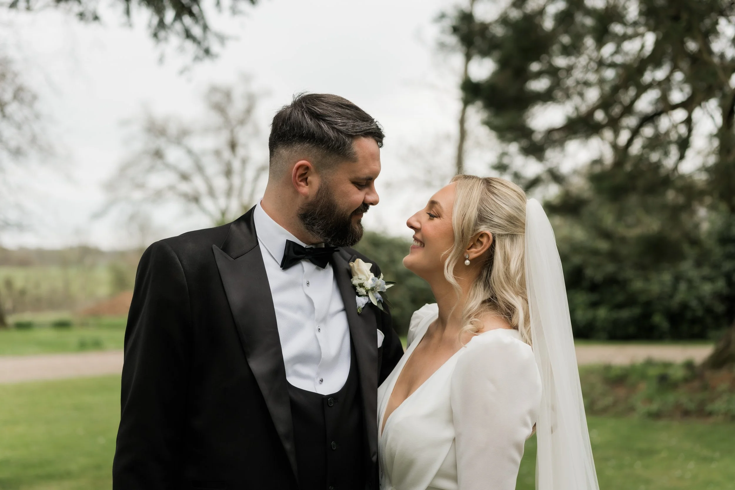 A bride and groom face each other outdoors, smiling, with trees and a grassy landscape in the background.