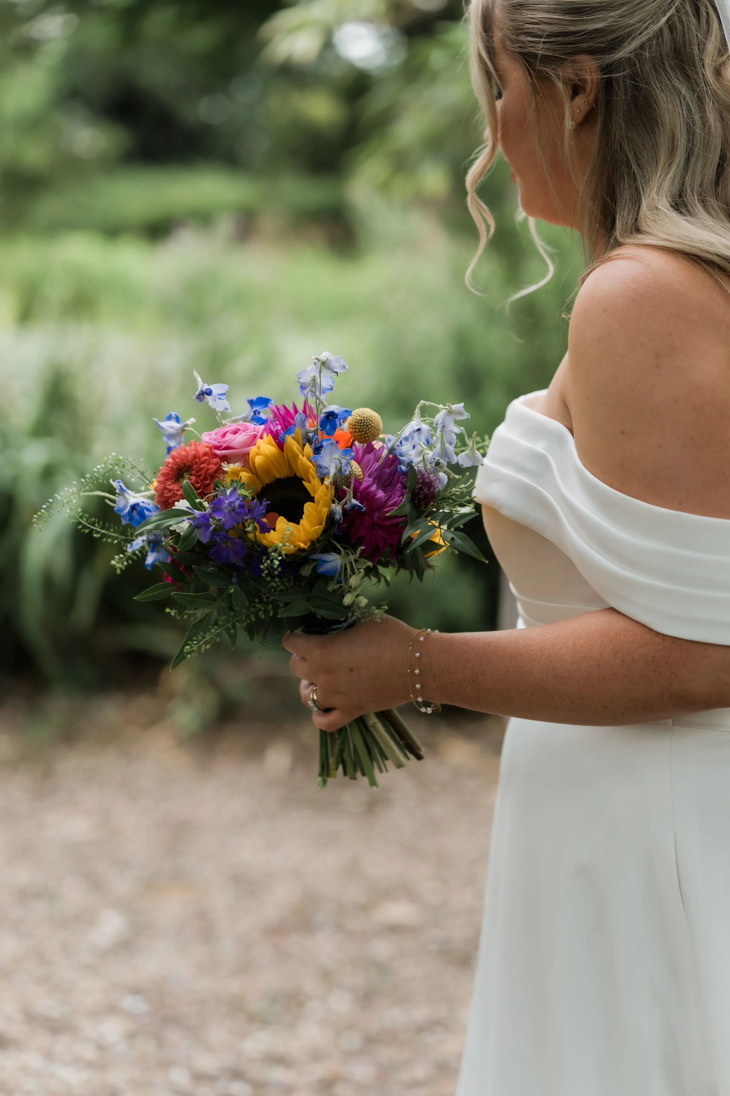 A woman in a white off-the-shoulder wedding dress holding a colorful bouquet of flowers, outdoors with a blurred green natural background.