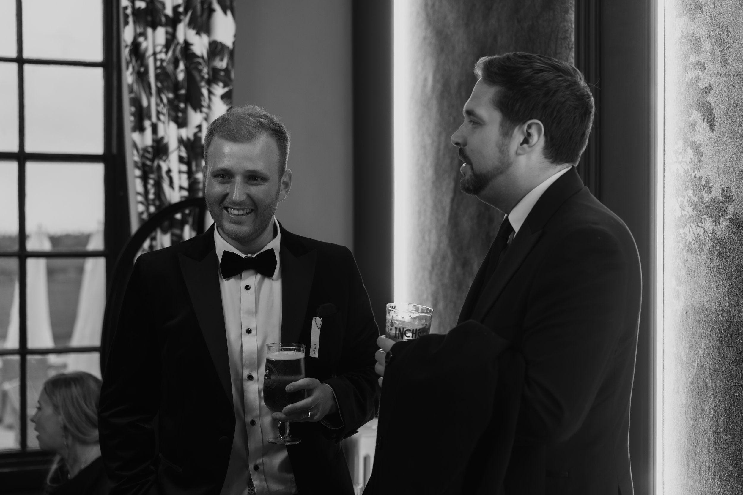 Two men in tuxedos with drinks, chatting at an indoor event, one smiling and the other listening, decorated interior with large windows and curtains.