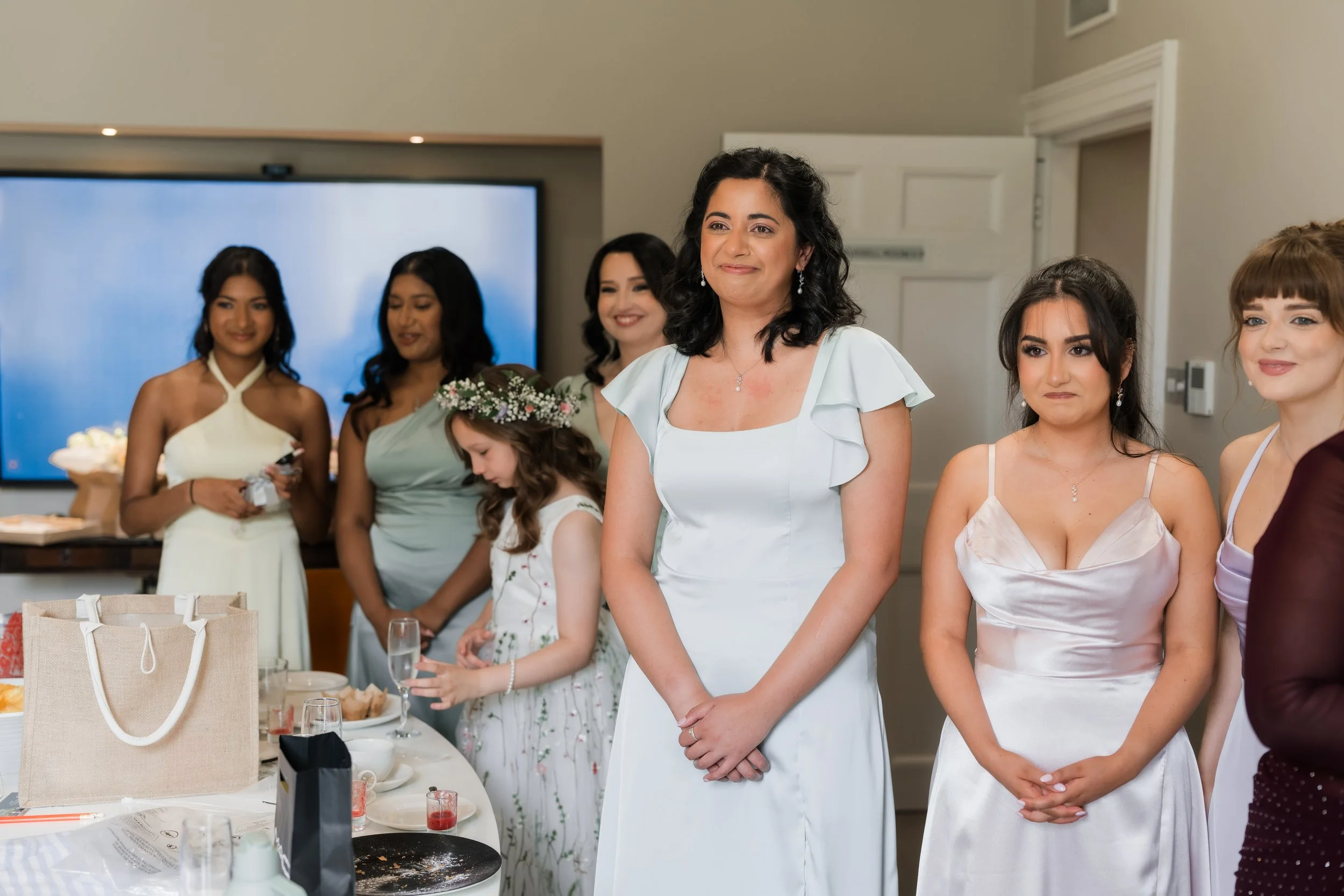 A group of women and a young girl, dressed in elegant dresses, at a celebration or gathering in a room with a large screen in the background.